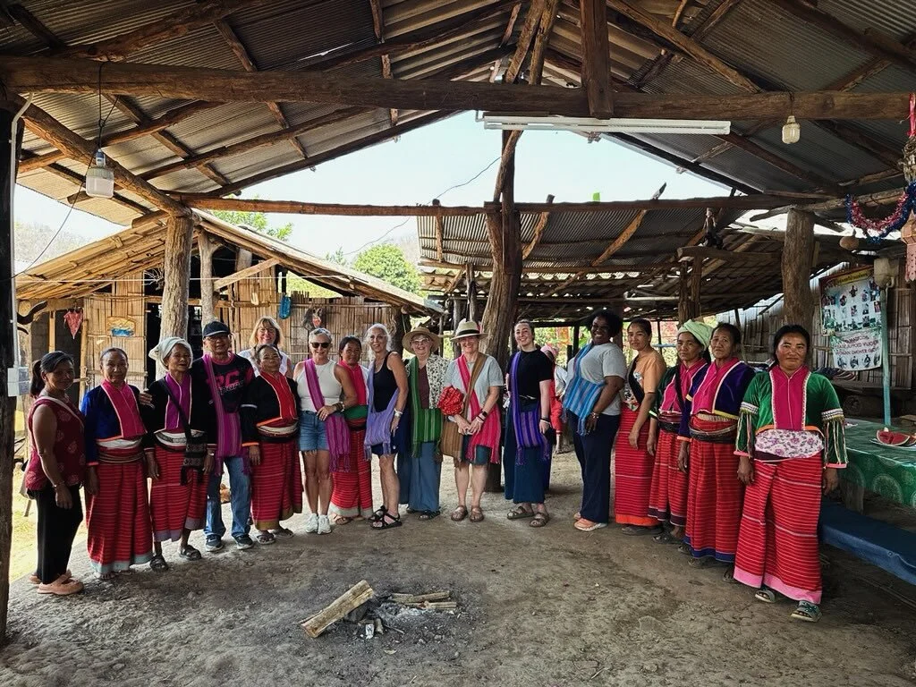 Group of women and visitors standing inside a rustic wooden structure, some women wearing colorful traditional clothing, with a thatched roof and bamboo walls, in a rural setting.