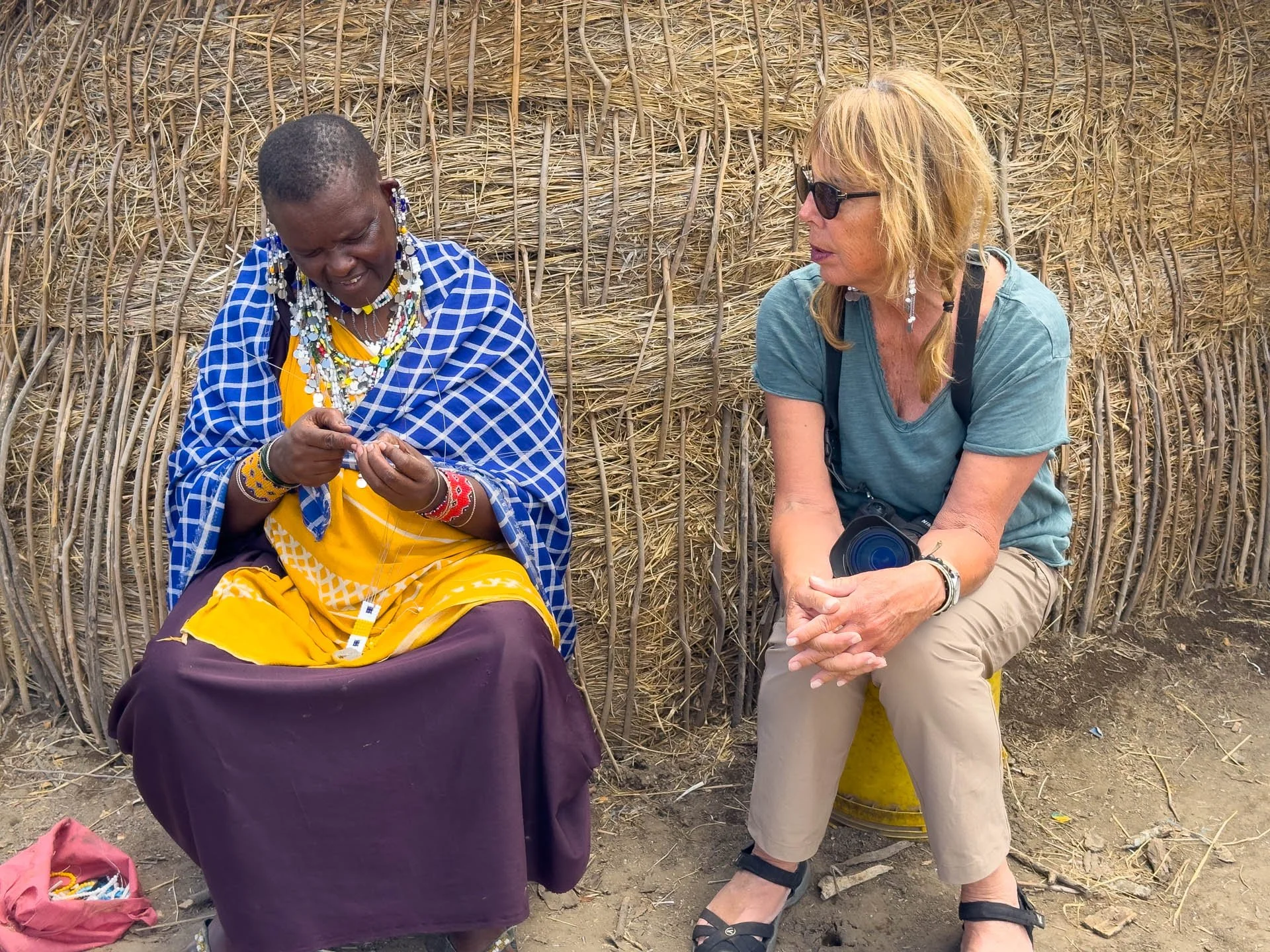 Caravan Gallery in La Conner, WA - Owner Linda watching a woman dressed in traditional Maasai attire sitting on a stool, sewing beaded jewelry, while a woman in casual clothing and sunglasses sits on a yellow stool nearby