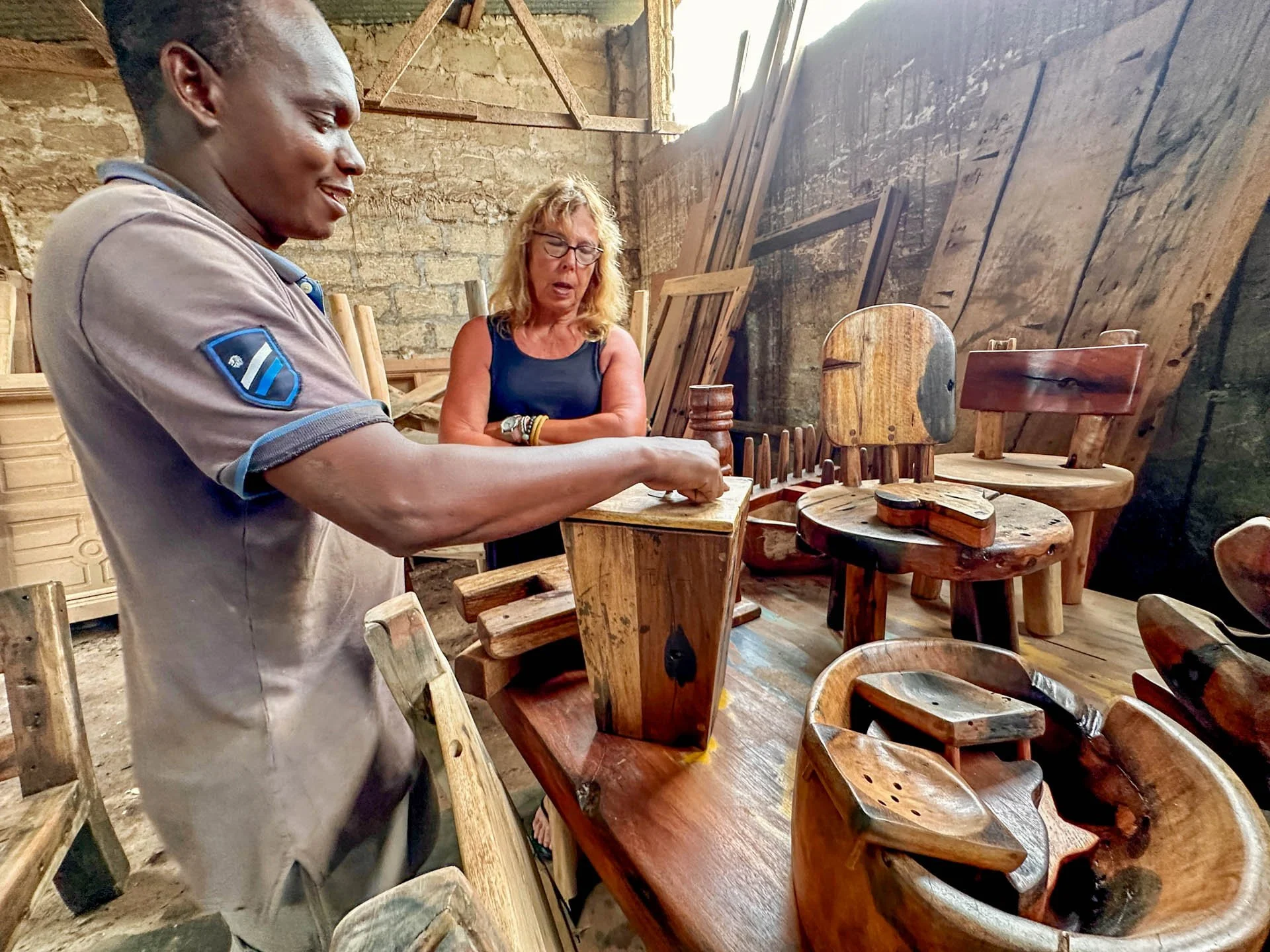 Caravan Gallery in La Conner, WA - Owner Linda in Zanzibar with a man making wooden handcrafted items on display in a rustic workshop with stone walls and wooden beams.