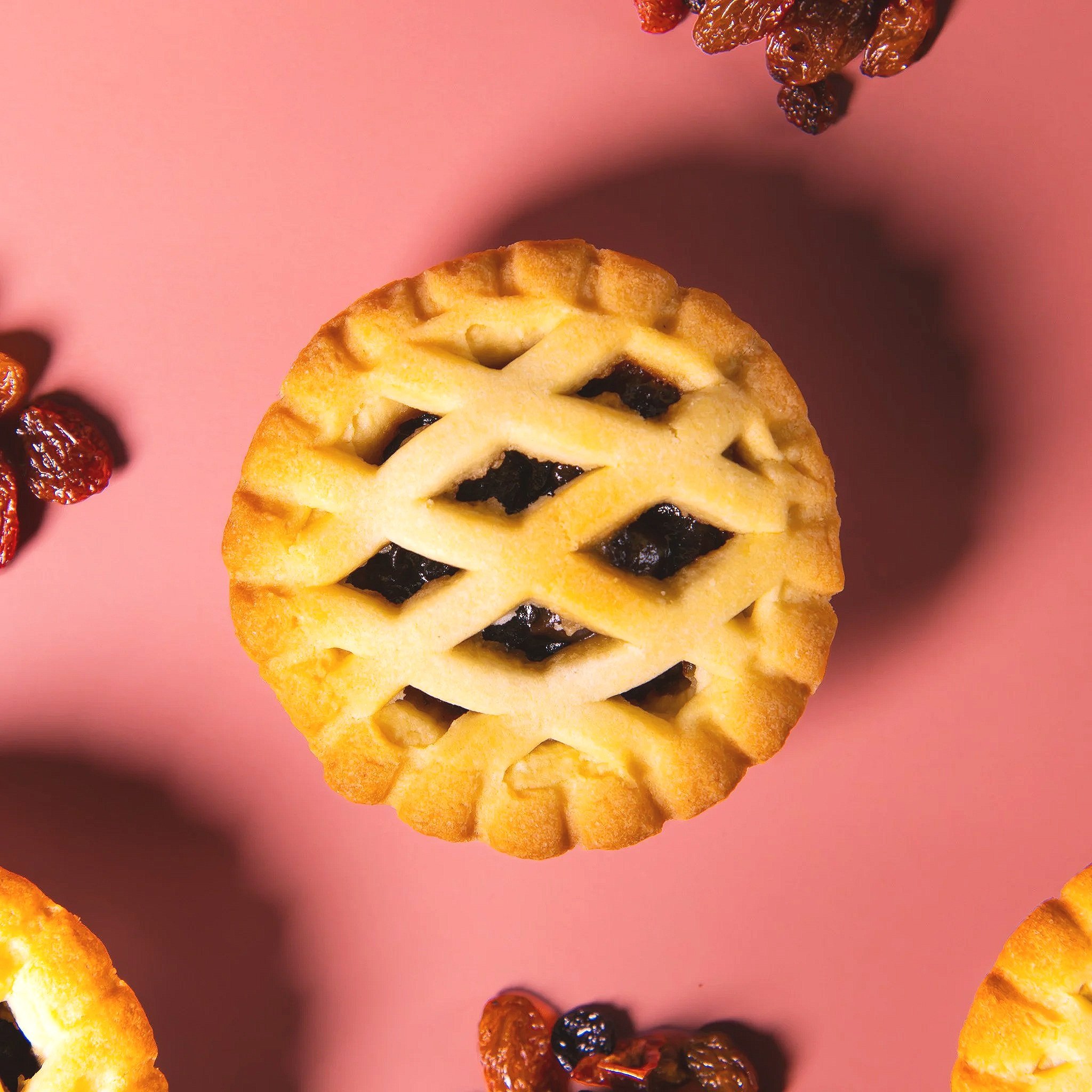 Top view of a freshly baked lattice-topped fruit tart on pink background, with scattered dried fruits around.