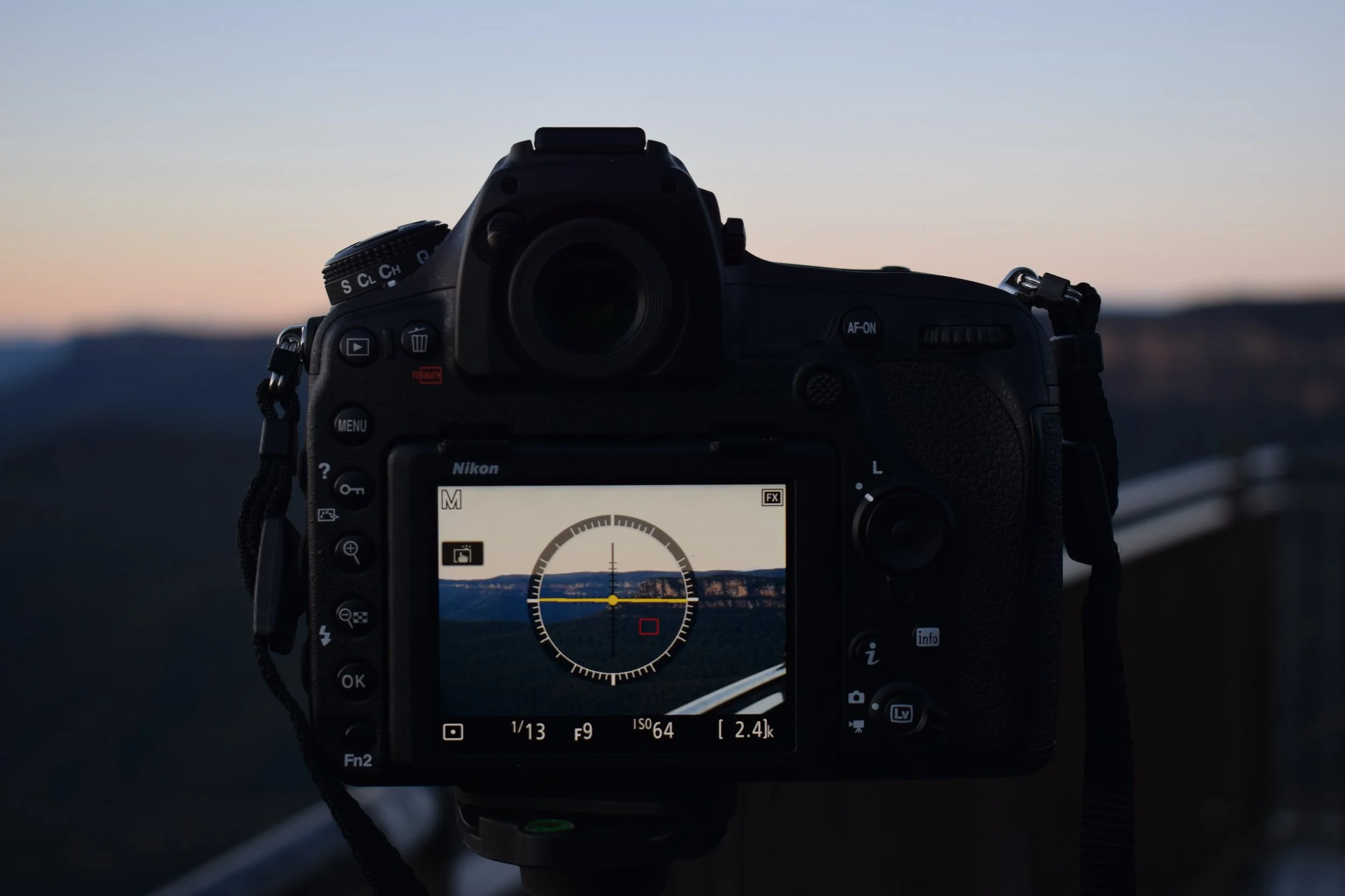 A digital camera positioned on a tripod capturing a landscape during sunset, with the camera's LCD screen displaying the scene of mountains and a road.