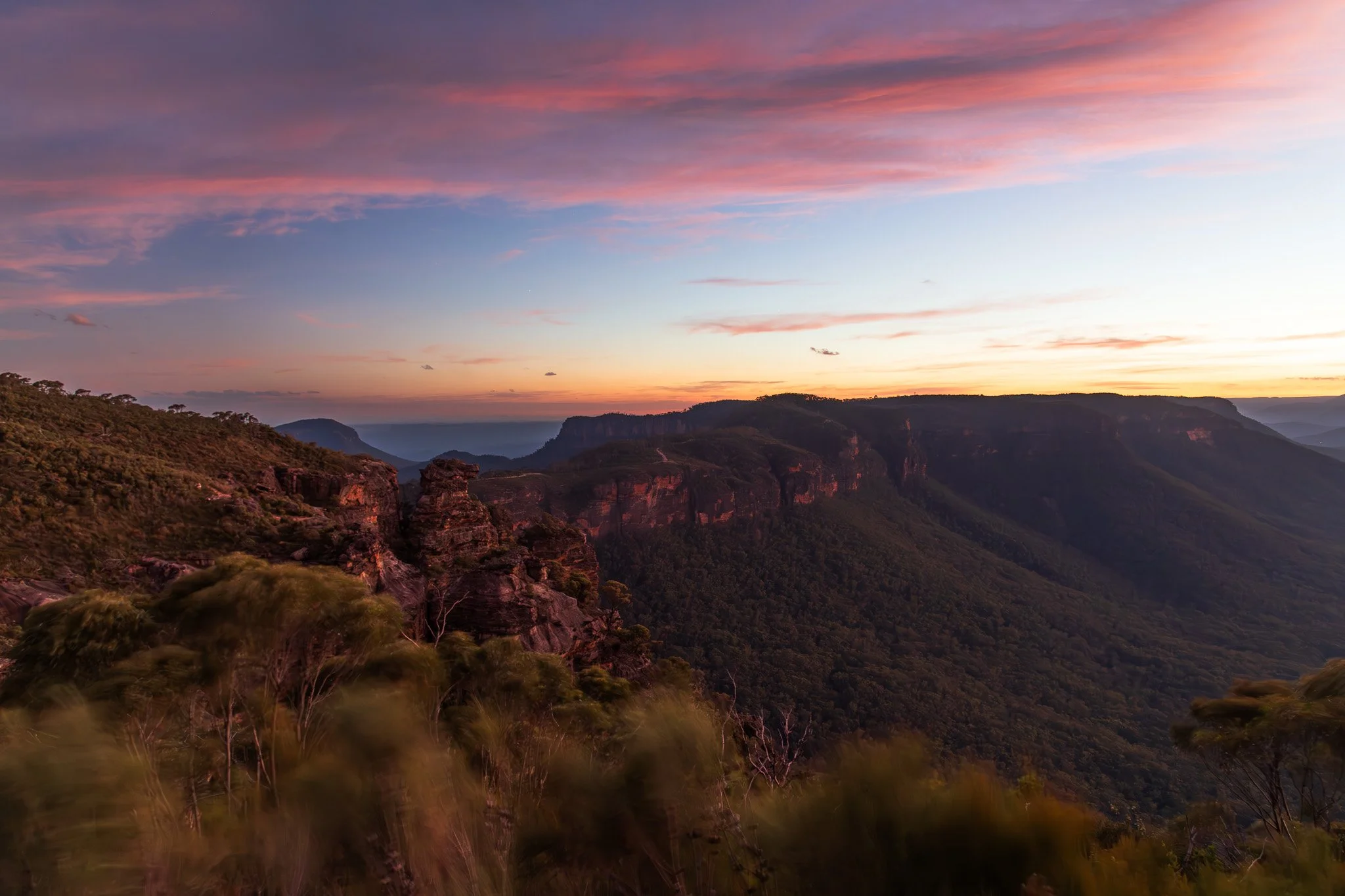 A scenic view of a mountainous landscape at sunset with layered hills and a colorful sky with scattered clouds.