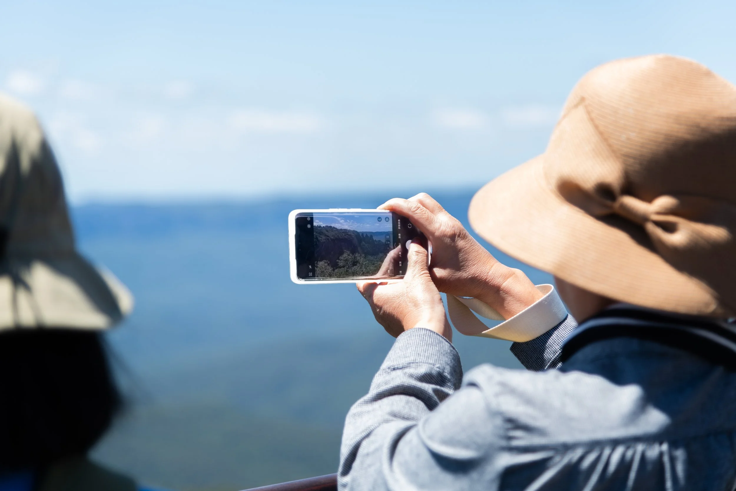 Person wearing a wide-brimmed hat and gray jacket taking a photo of a landscape with a smartphone, with plans of water and mountains in the background.