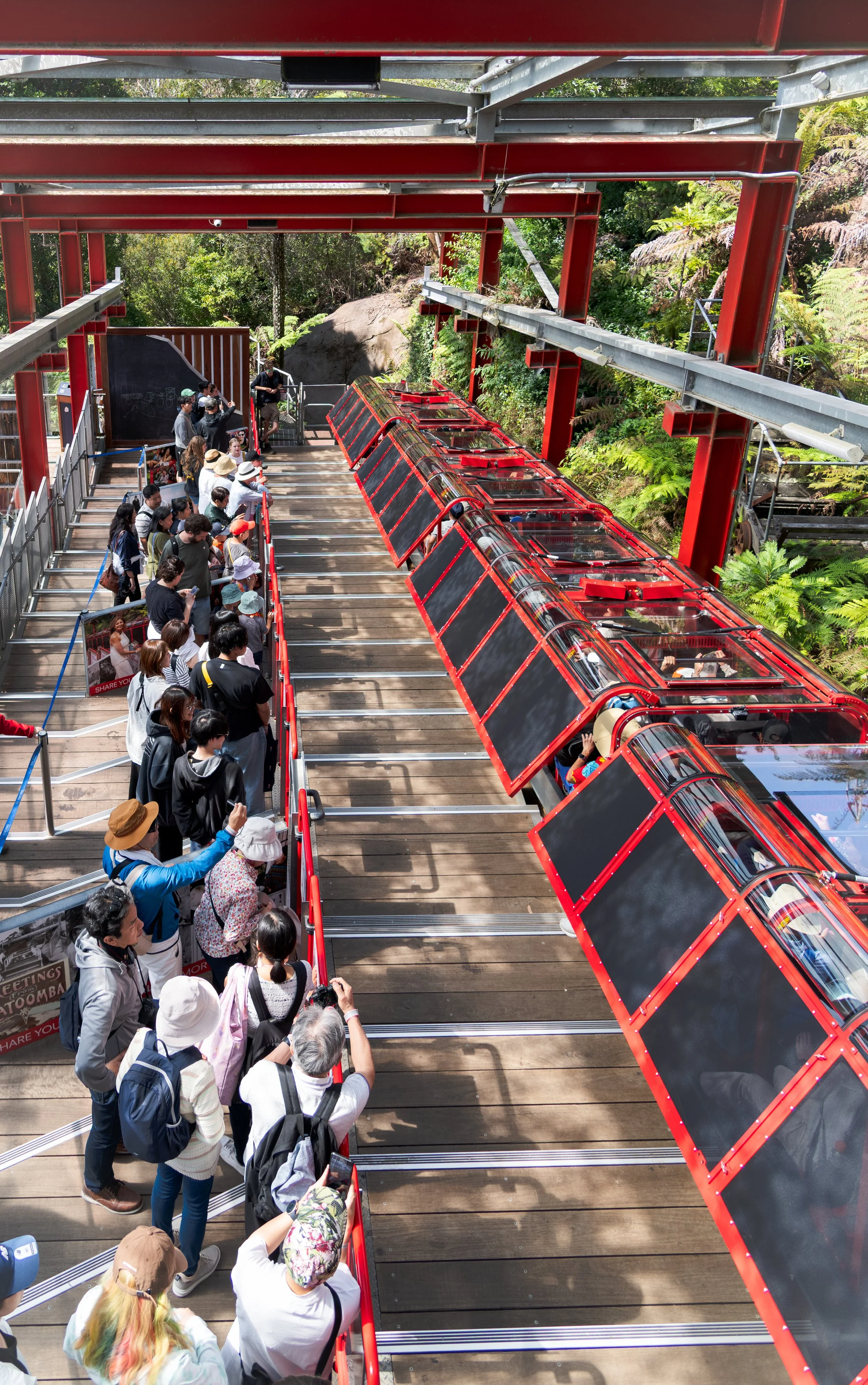 People lining up on a wooden platform to board a red, enclosed cable car or gondola lift with glass windows, surrounded by lush green trees and foliage.