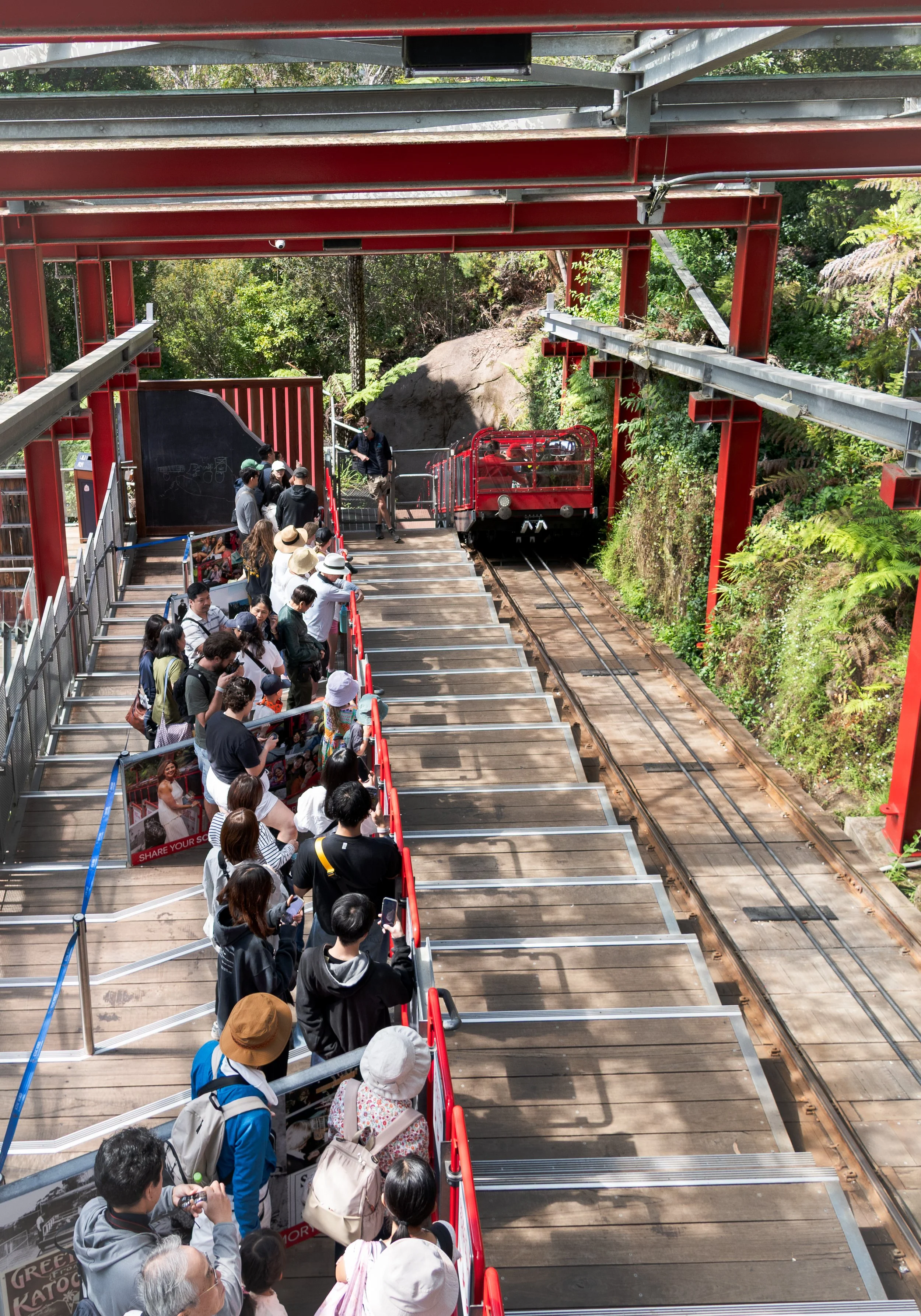 Line of people waiting to ride a narrow-gauge train on a track through a green, forested area.