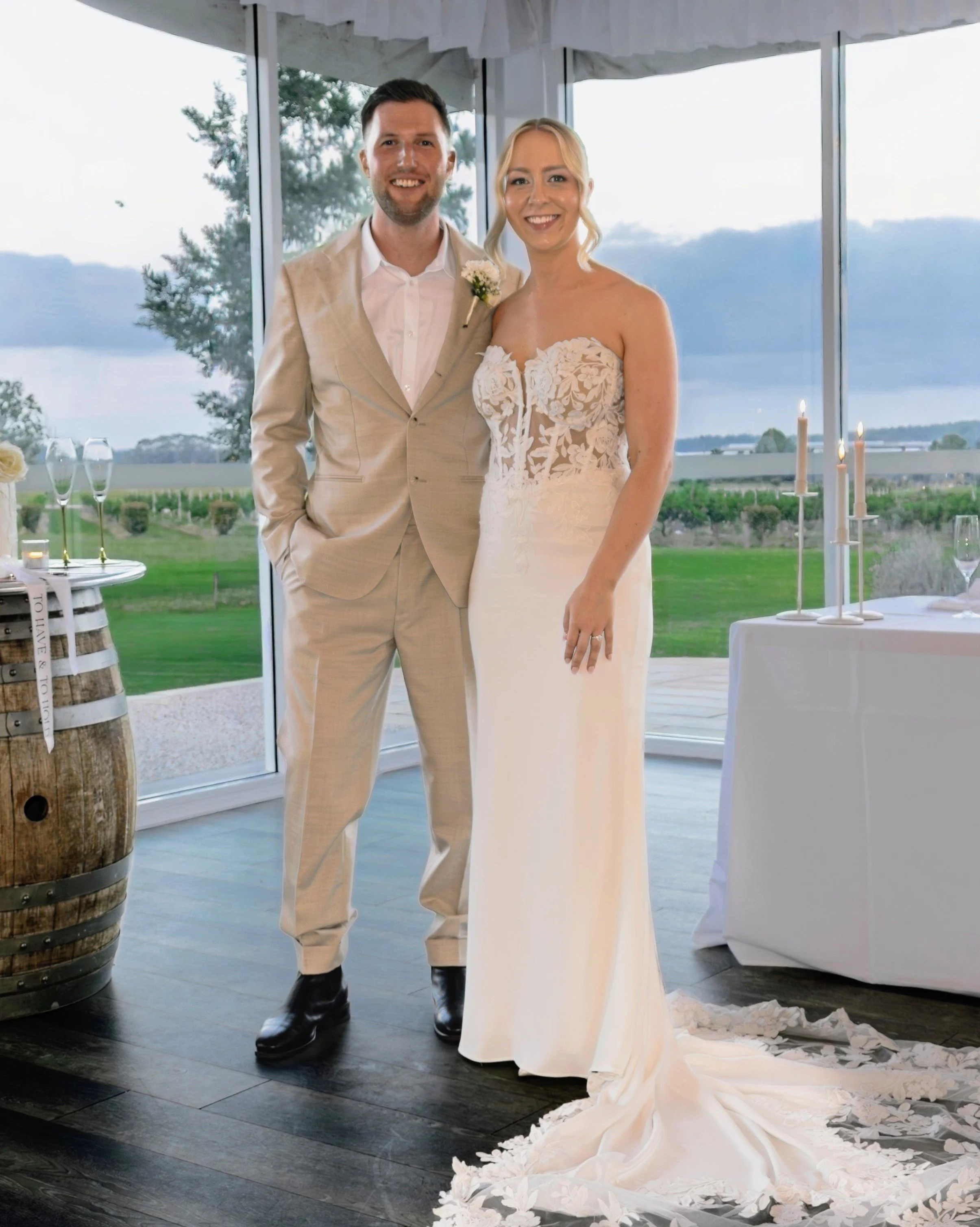 A newlywed couple standing indoors near a window with a scenic outdoor view, the groom in a beige suit and the bride in an off-shoulder white wedding gown with lace details, smiling at the camera.