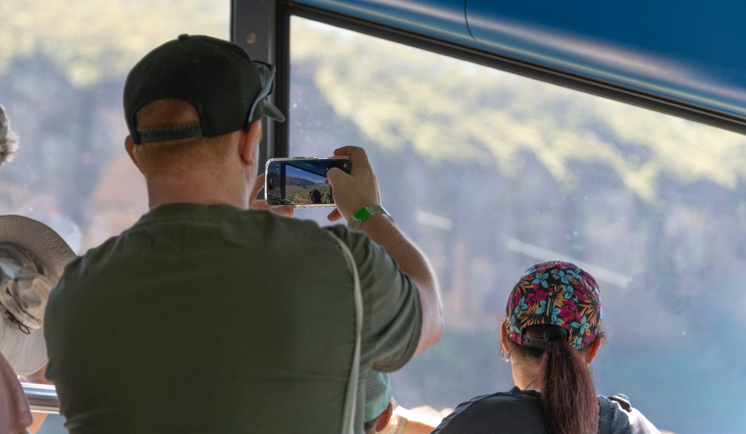 A person with a baseball cap and sunglasses taking a photo with their phone on a bus, next to a woman wearing a floral cap.