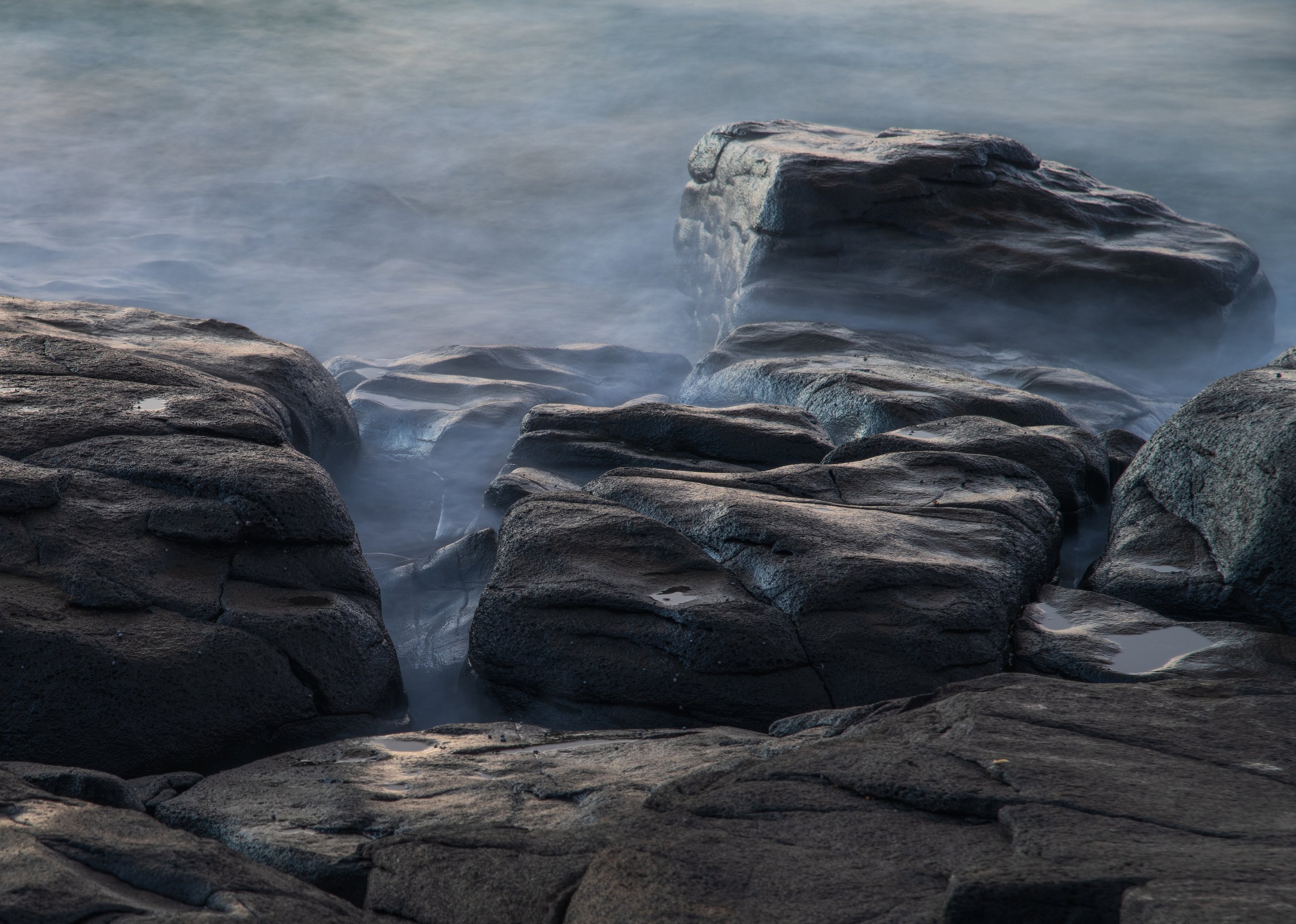 A rocky shoreline with water gently washing over dark rocks, creating a misty effect around the stones.