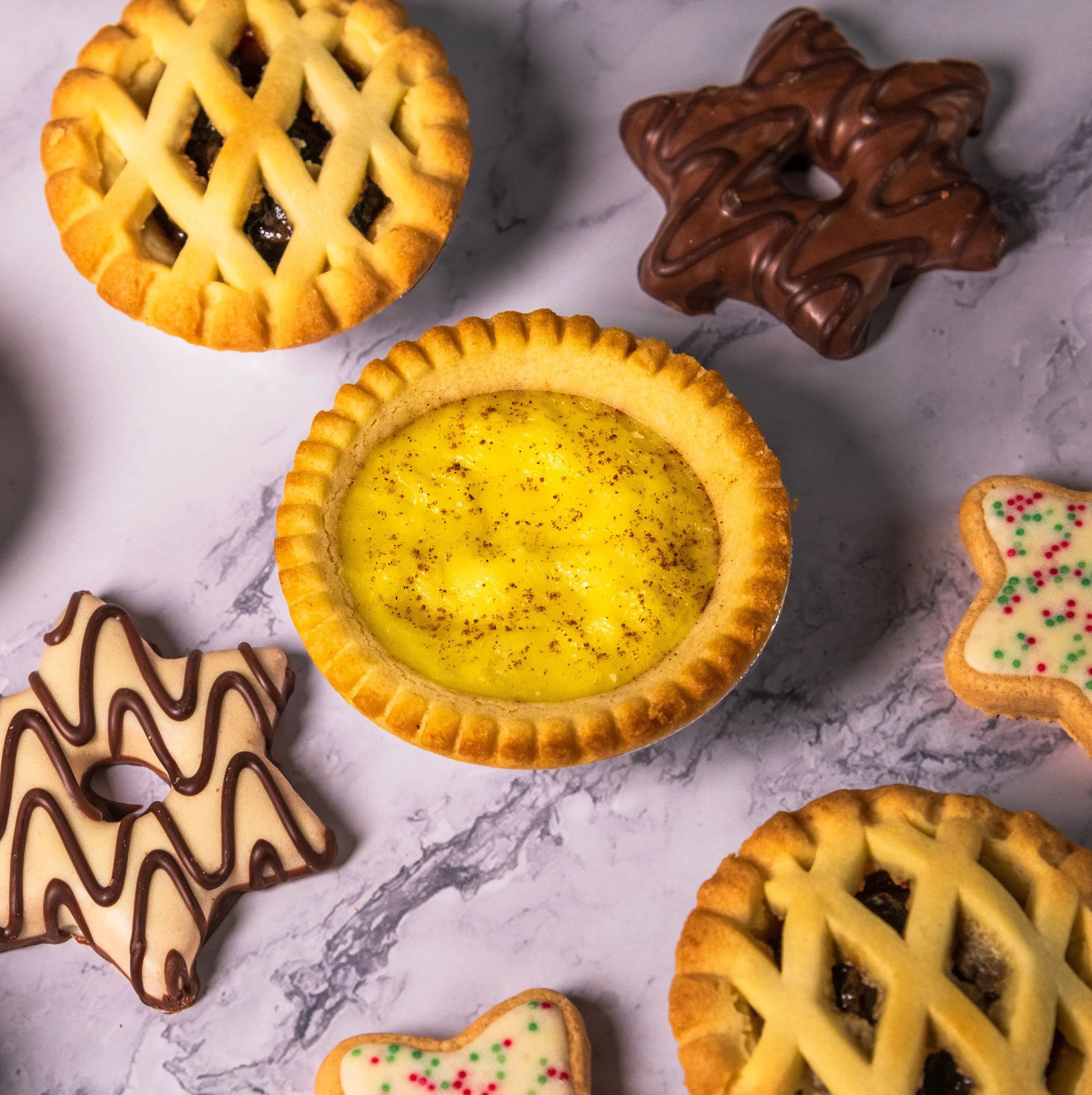 Assortment of Christmas-themed cookies, including star-shaped cookies with white icing and green and red sprinkles, pie cookies with lattice crust, chocolate drizzled cookies, and a lemon tart with a golden crust and sprinkled with pepper, arranged o