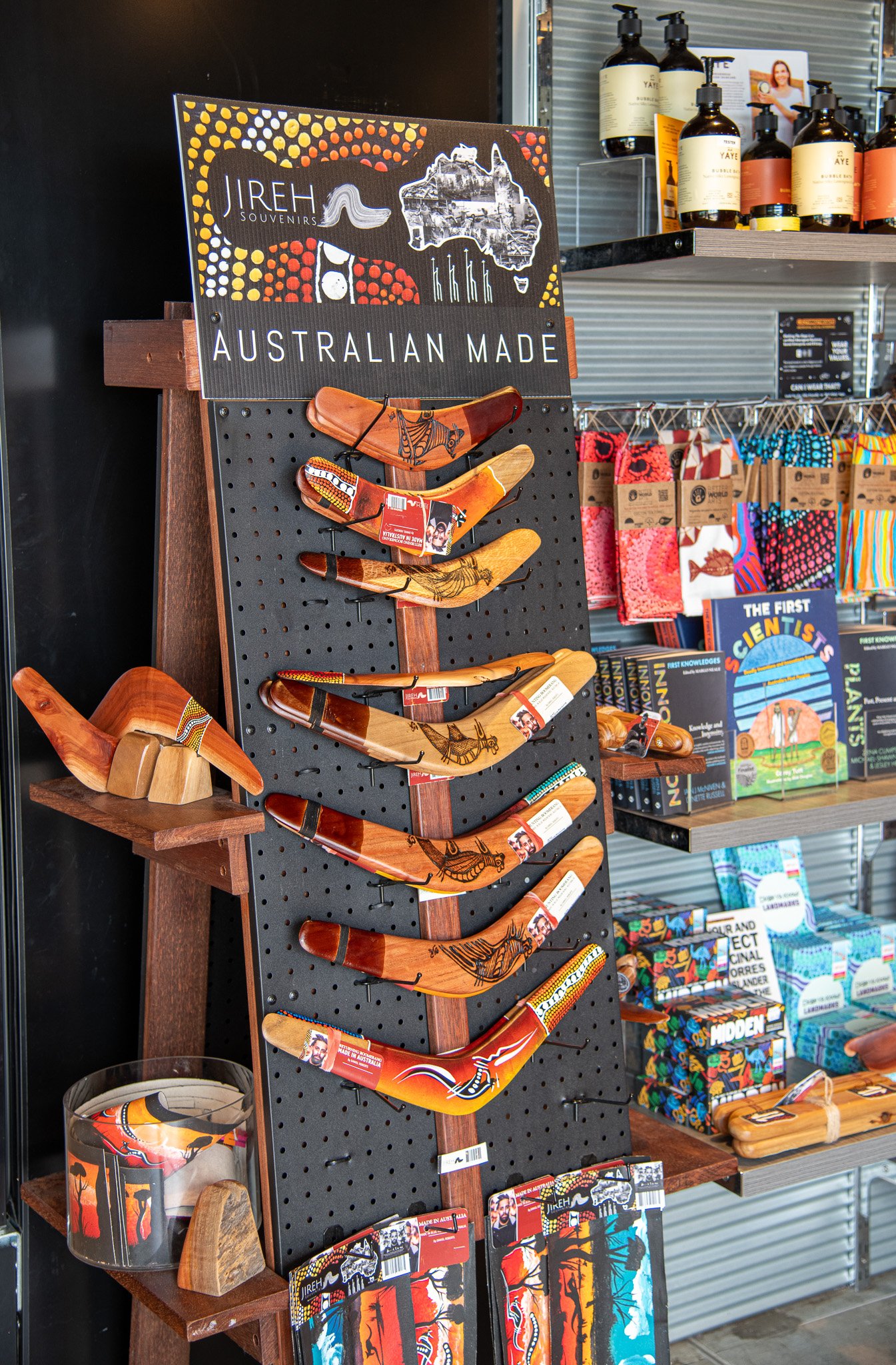 Display of Aboriginal boomerangs and souvenirs at a store called Jireh Souvenirs, with shelves of bottles and books in the background.