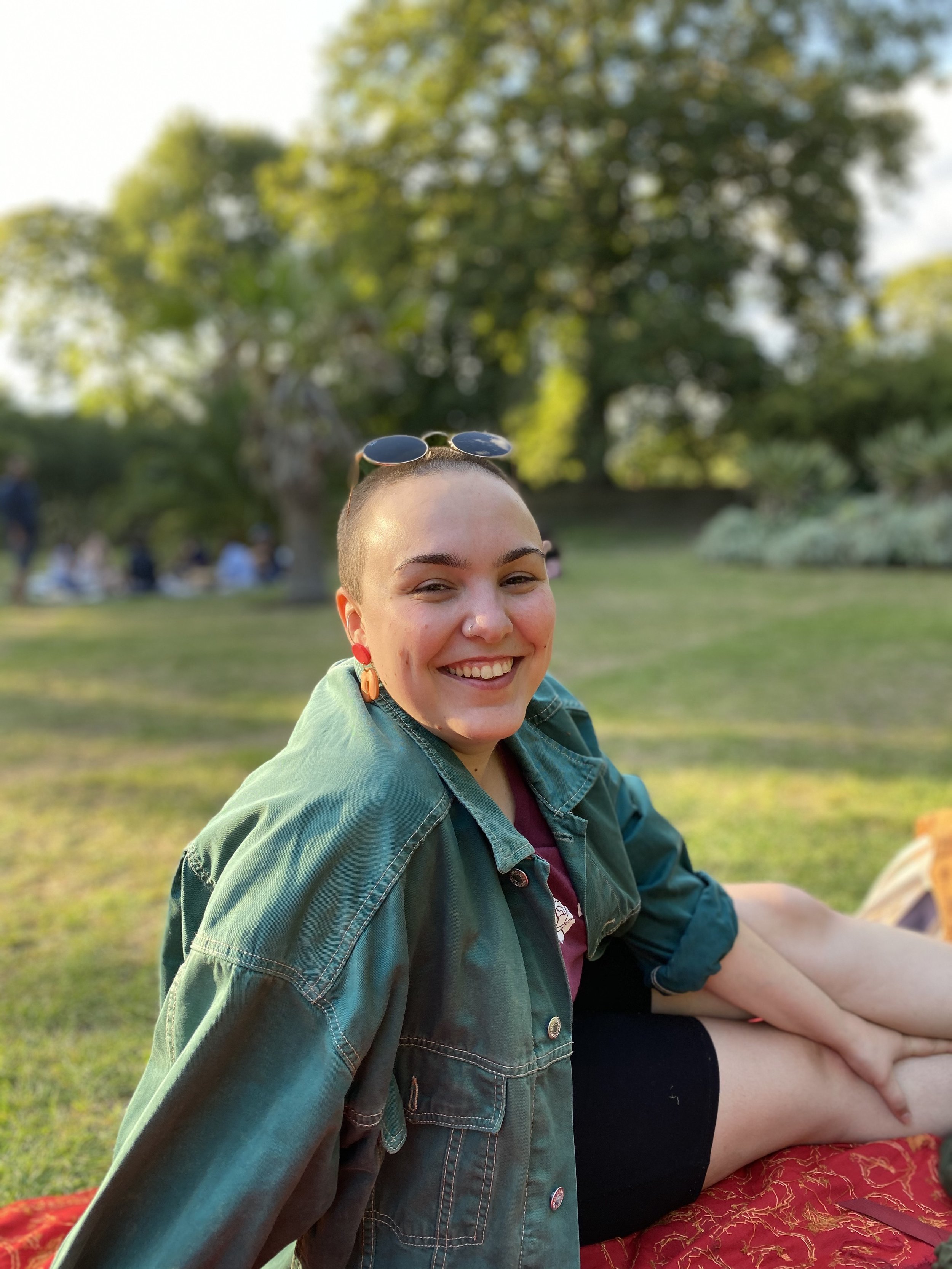 Smiling young woman with short hair, wearing sunglasses on her head, earrings, a teal jacket, and a maroon shirt, sitting on the grass in a park during sunny weather.