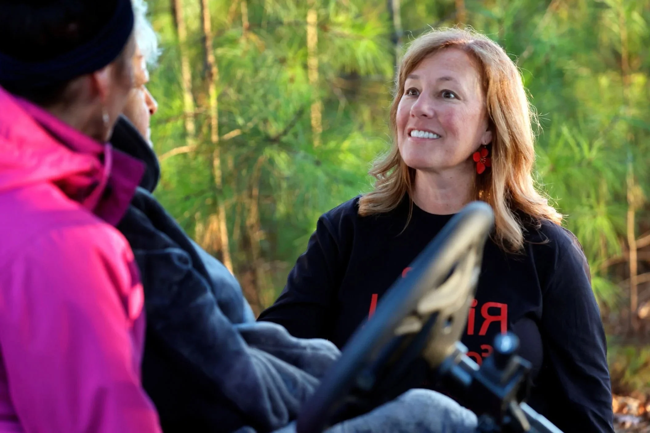 A woman with shoulder-length blonde hair, wearing earrings and a black T-shirt, smiling and speaking to two other women in a forested outdoor setting.