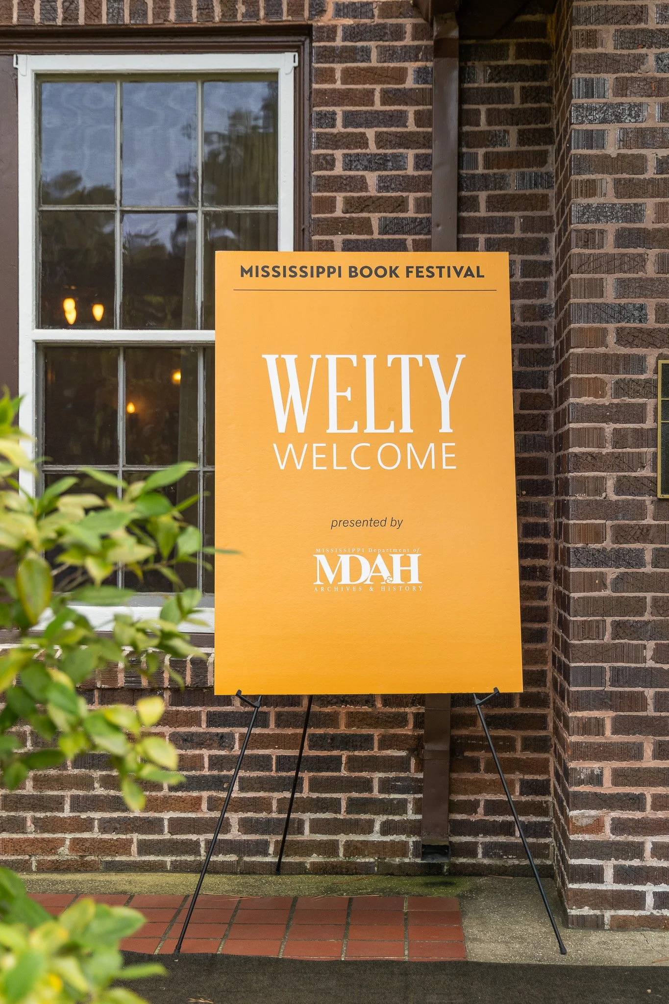 Orange sign with black and white text welcoming attendees to the Mississippi Book Festival, presented by the Mississippi Department of Archives & History, placed outside near a brick building with a window.