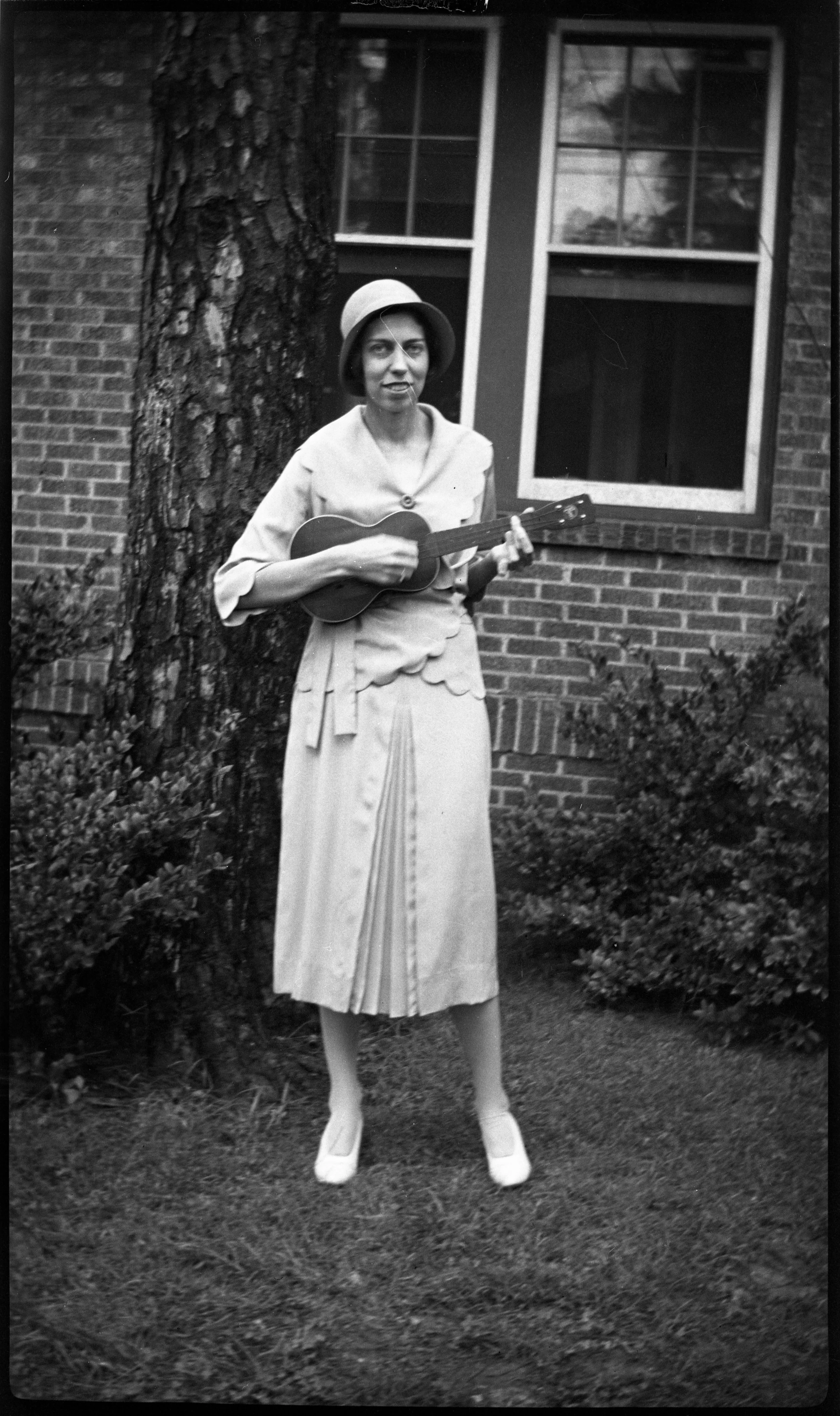 A woman wearing a hat and a long dress, standing outdoors near a tree, playing a ukulele, in front of a brick house with an open window.