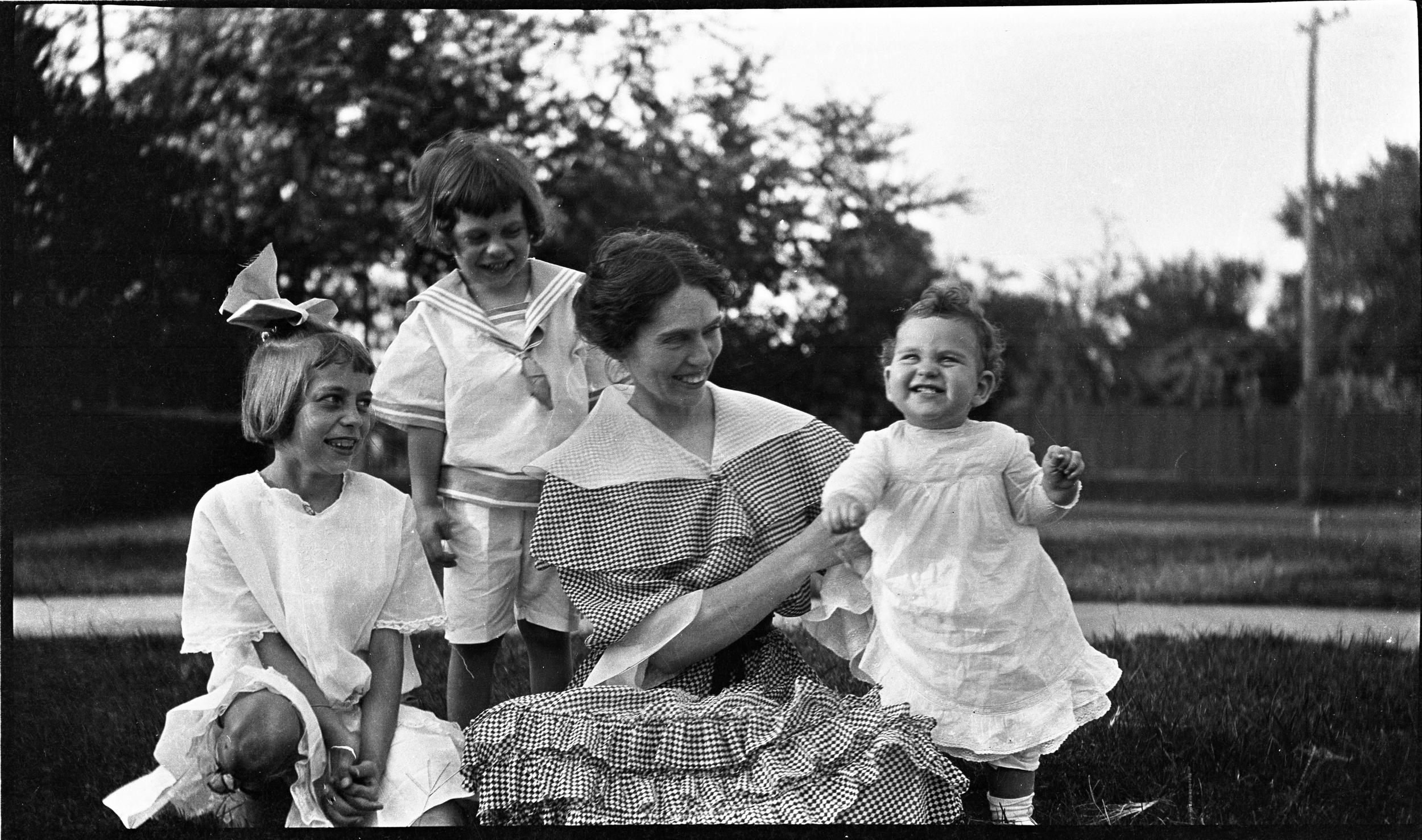 Black and white photo of a woman and three young girls outside in a park or yard, smiling and playing together. The woman is holding a small girl in a white dress, while the others are watching.