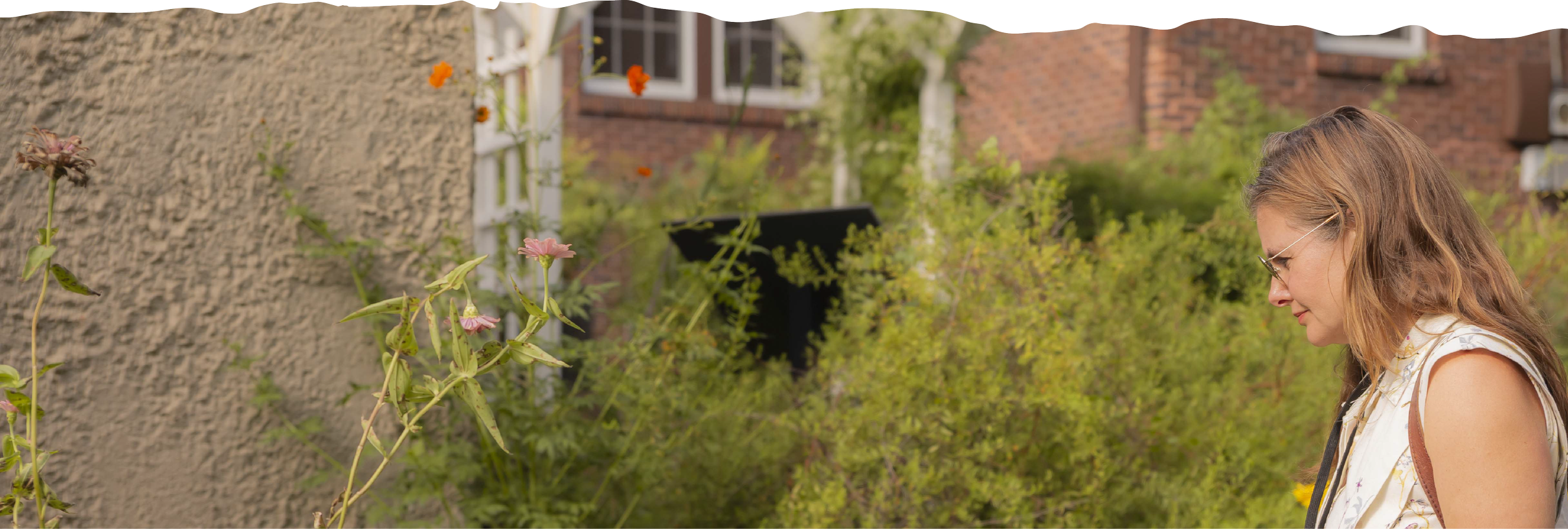 A woman with long wavy hair and glasses standing in a garden with various plants and flowers, looking down thoughtfully.