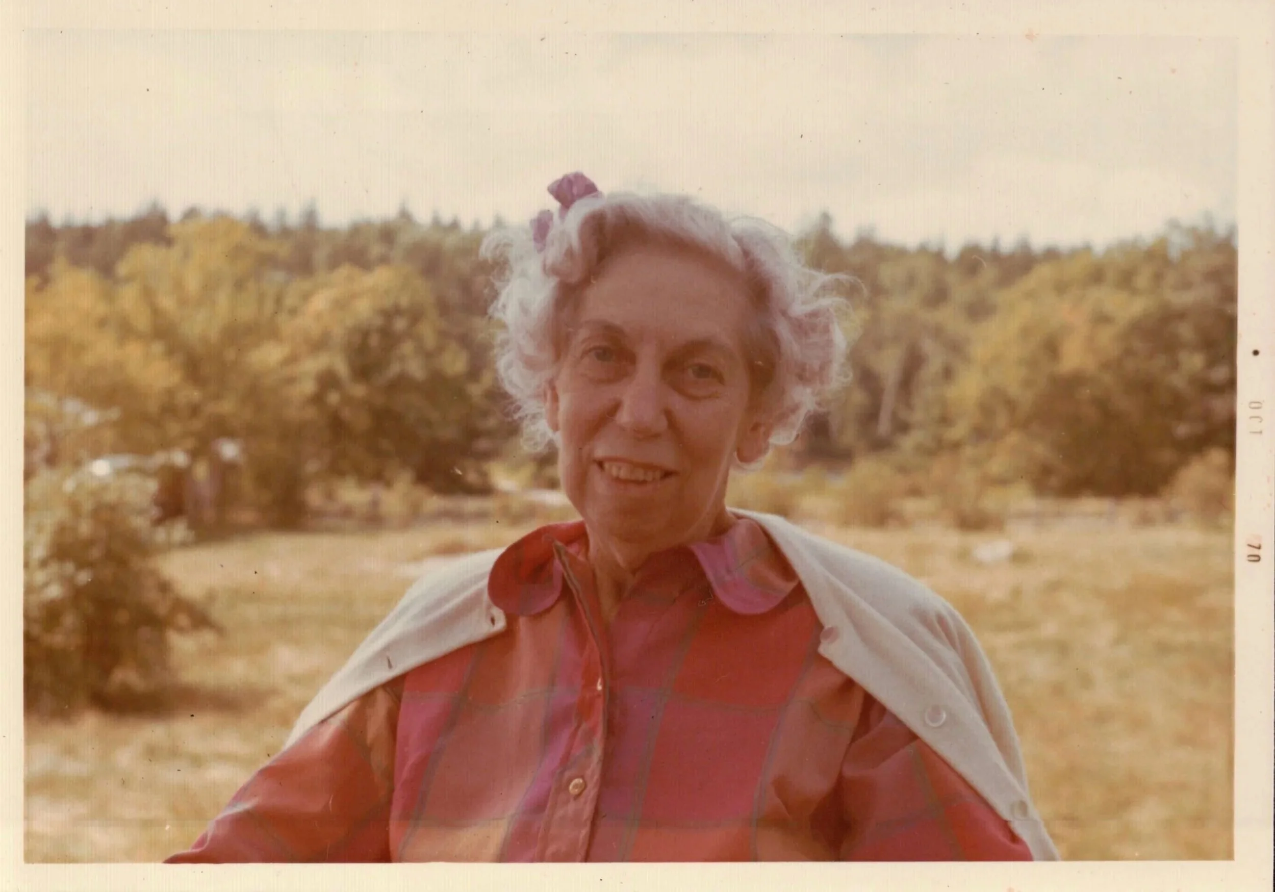 An elderly woman outdoors with a background of trees and open field, wearing a red plaid shirt and a light jacket draped over her shoulders, smiling at the camera.