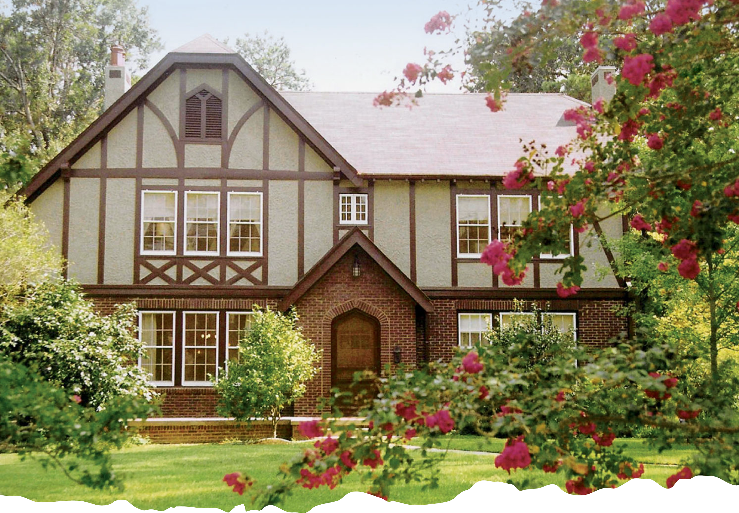 A large house with Tudor-style architecture, featuring a brick lower level, stucco upper level with decorative wood beams, and multiple windows, with a lush green lawn and pink flowering trees in the foreground.