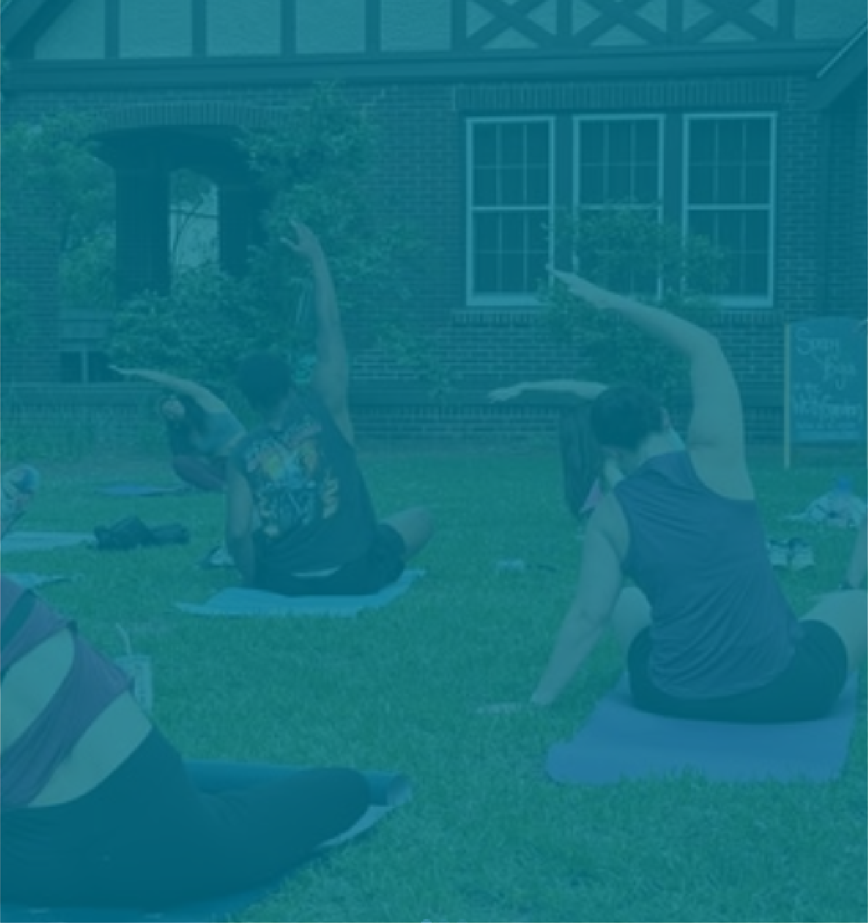 People sitting on yoga mats outdoors, practicing yoga or stretching in a grassy area outside a brick building with windows.