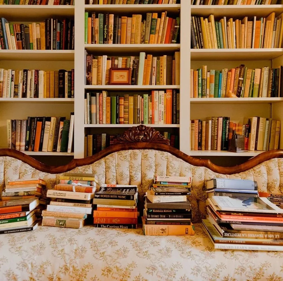 Bookshelves filled with numerous books above a vintage floral sofa with stacks of books on it.