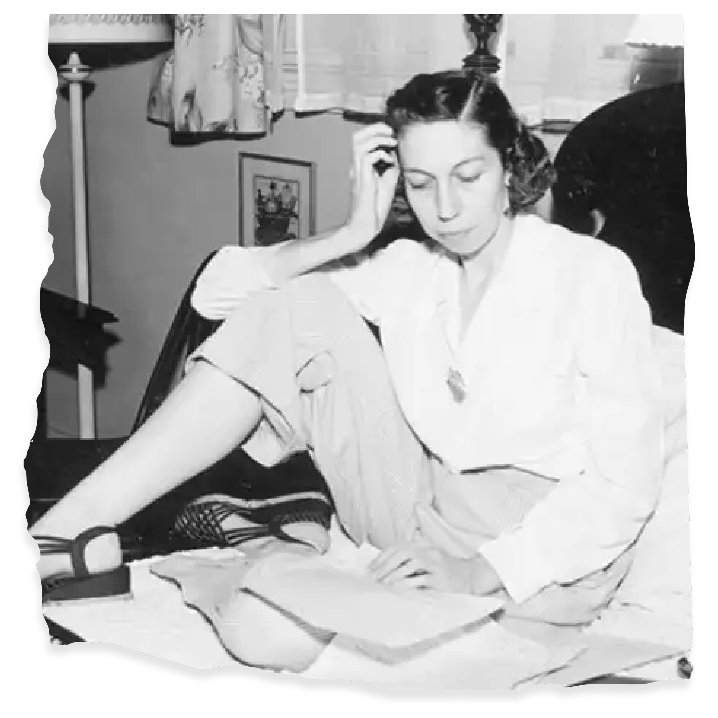 A black and white photo of a woman sitting on a bed, with her eyes closed and her hand on her head. She is wearing a light-colored shirt, shorts, and fishnet stockings. There are papers or a book on her lap. In the background, there is a bedside lamp, a framed picture on the wall, curtains, and a dark headboard.
