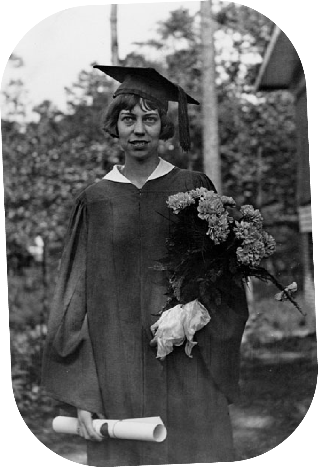 Black and white photo of a woman in a graduation cap and gown holding a bouquet of flowers and a diploma outdoors.