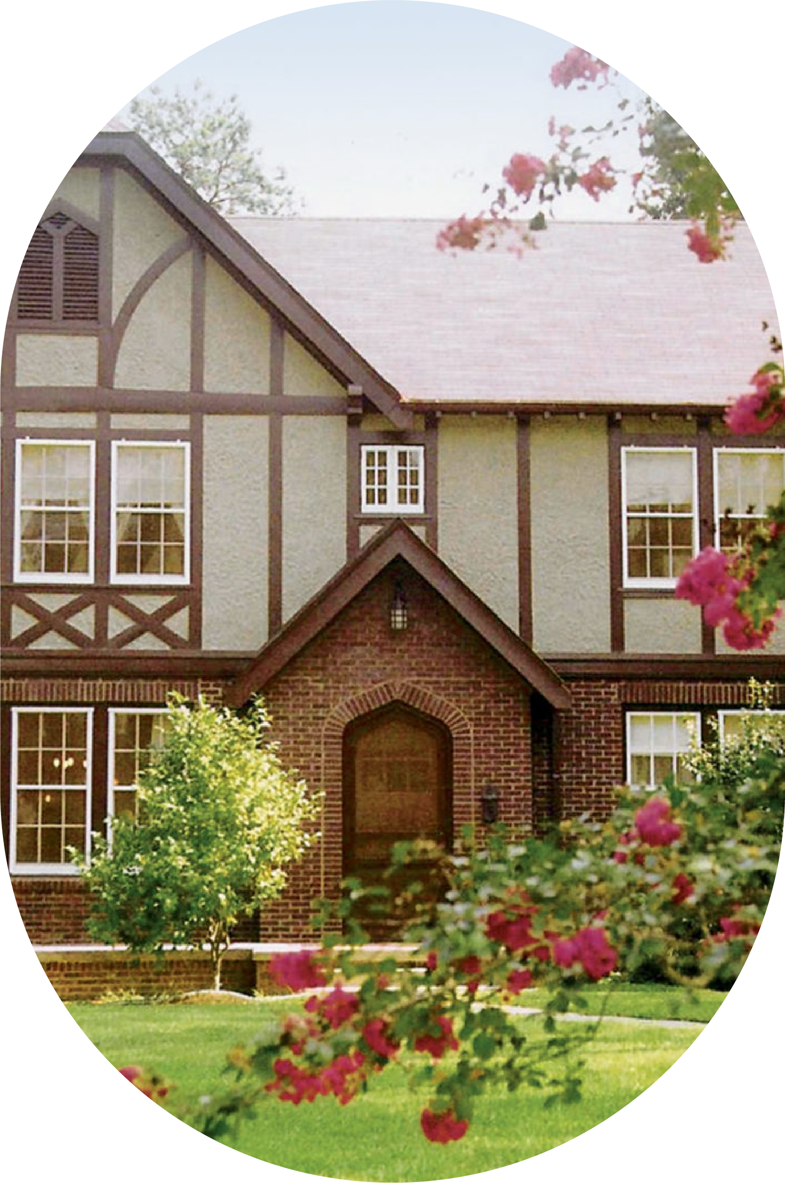A two-story house with a brick entrance and cream-colored siding with brown trim, surrounded by a green lawn and pink flowers.