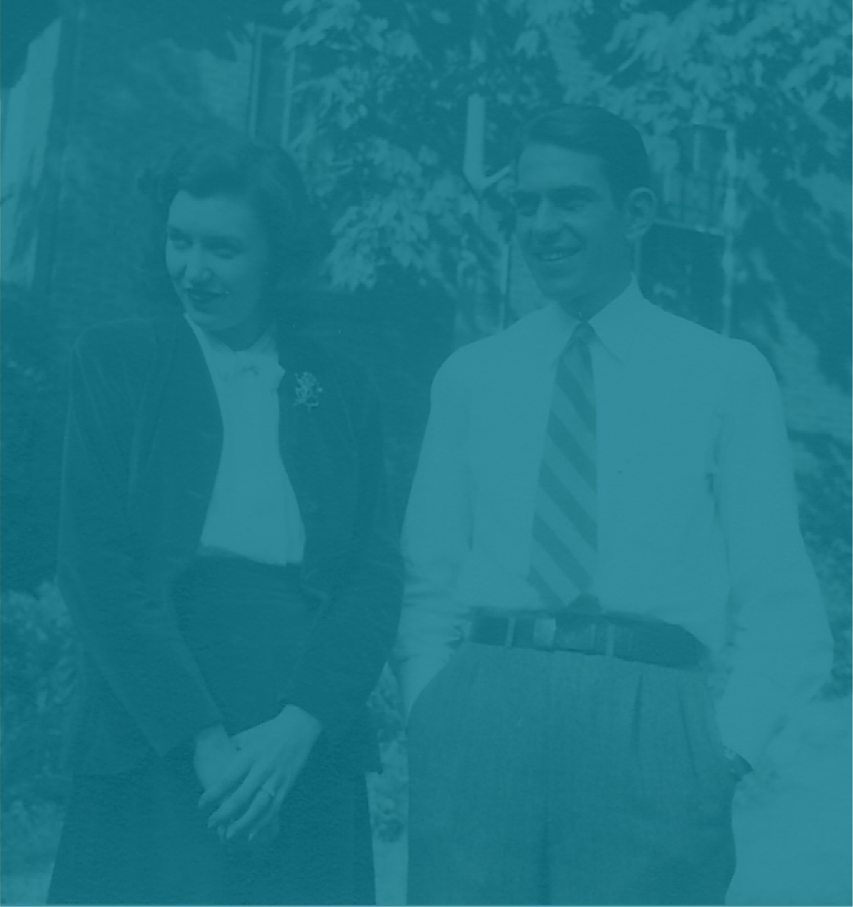 A woman and a man walking outdoors, smiling and talking, with trees and a house in the background.