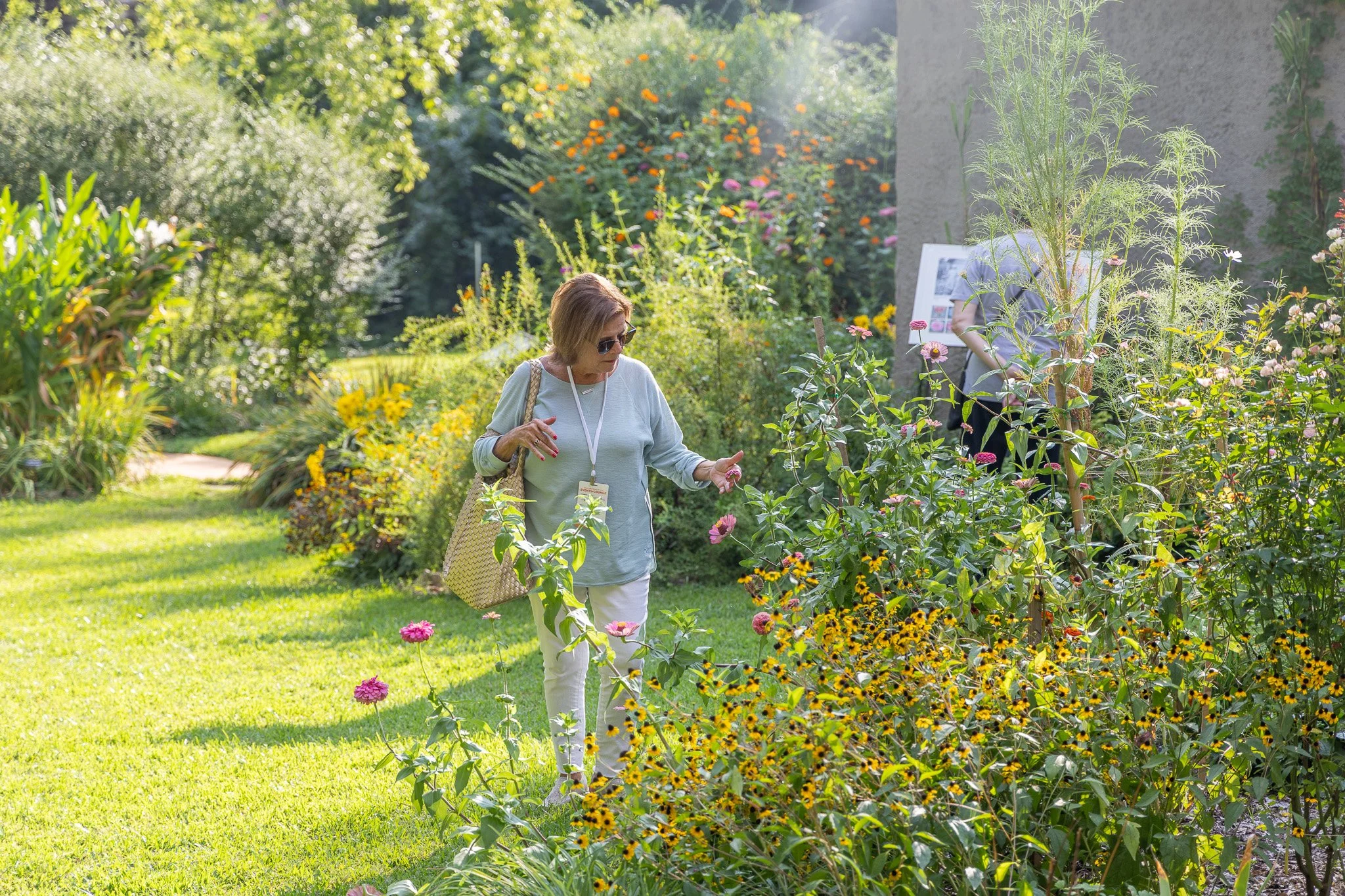 A woman with sunglasses and a beige bag walking through a lush garden filled with colorful flowers, trees, and plants, with a large wall or structure with informational displays behind her.