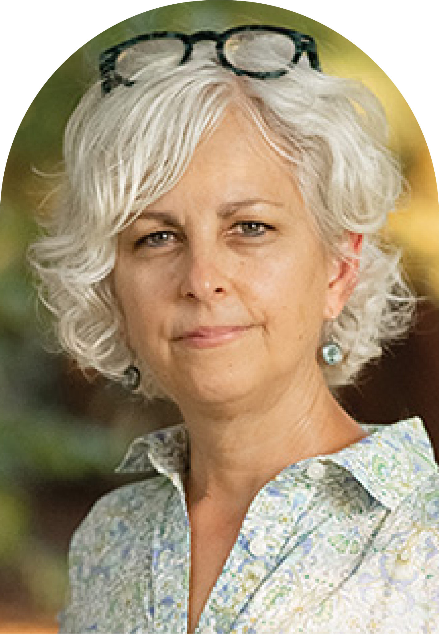A portrait of an Kate DiCamillo with curly gray hair, wearing glasses on her head and earrings with a white patterned shirt, outdoors with a blurred green and yellow background.