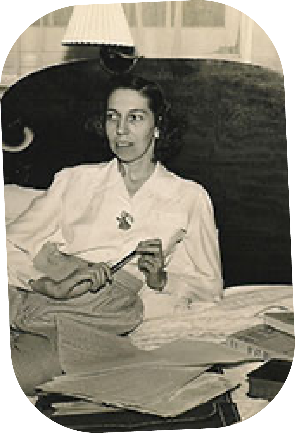 A woman sitting at a desk, holding a pen near some papers, with a lamp in the background.