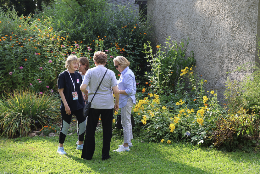 Four women stand in a garden with various flowers and plants, conversing and looking at something.