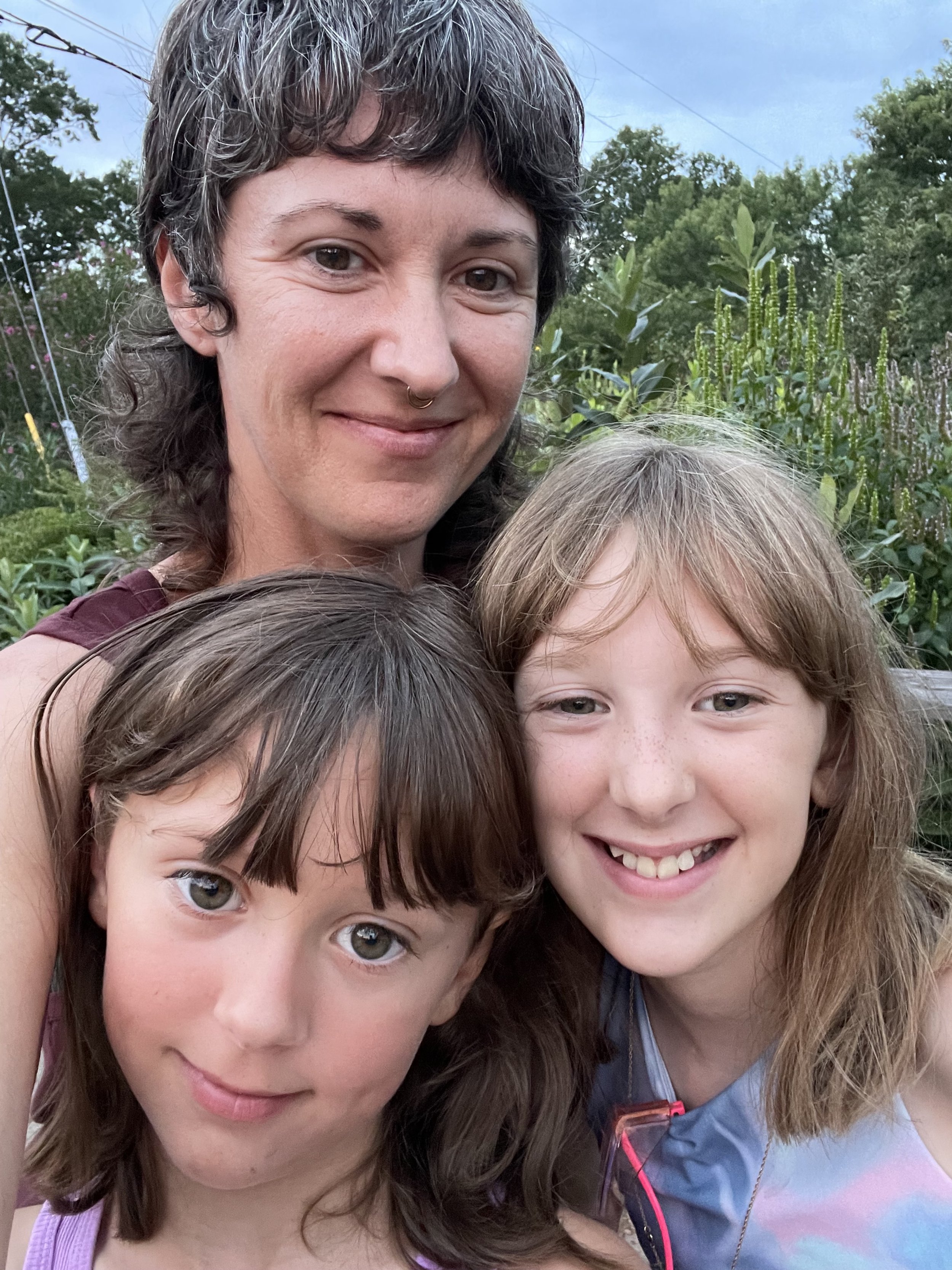 Three females smiling outdoors in front of greenery. An adult woman with dark wavy hair, a septum piercing, and two young girls with light-colored hair and blue eyes, one with straight hair and the other with slightly wavy hair.
