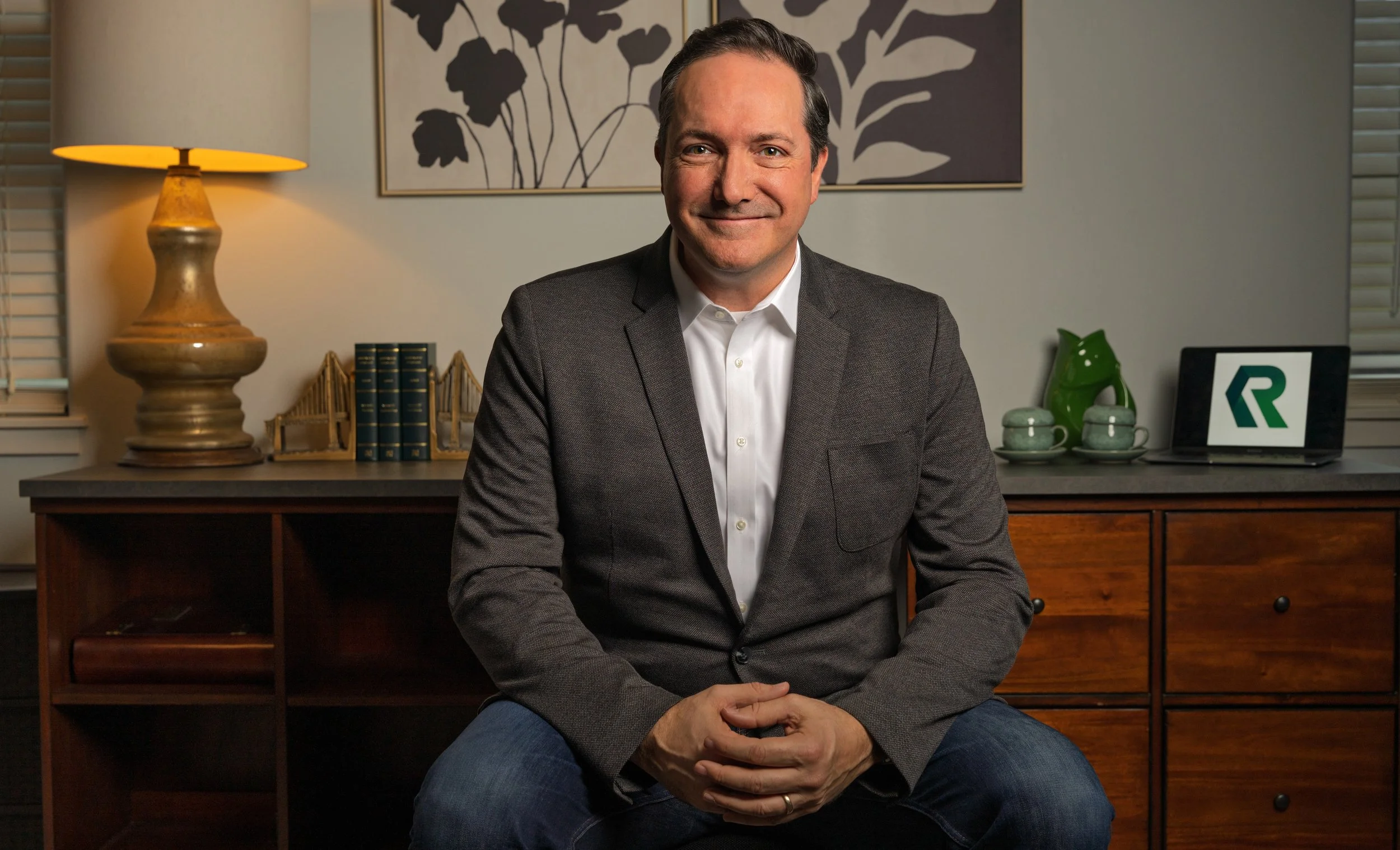 A man sitting in a room with a wooden desk, a lamp, books, a green dragon figurine, cups, and a laptop with a geometric logo, smiling at the camera.