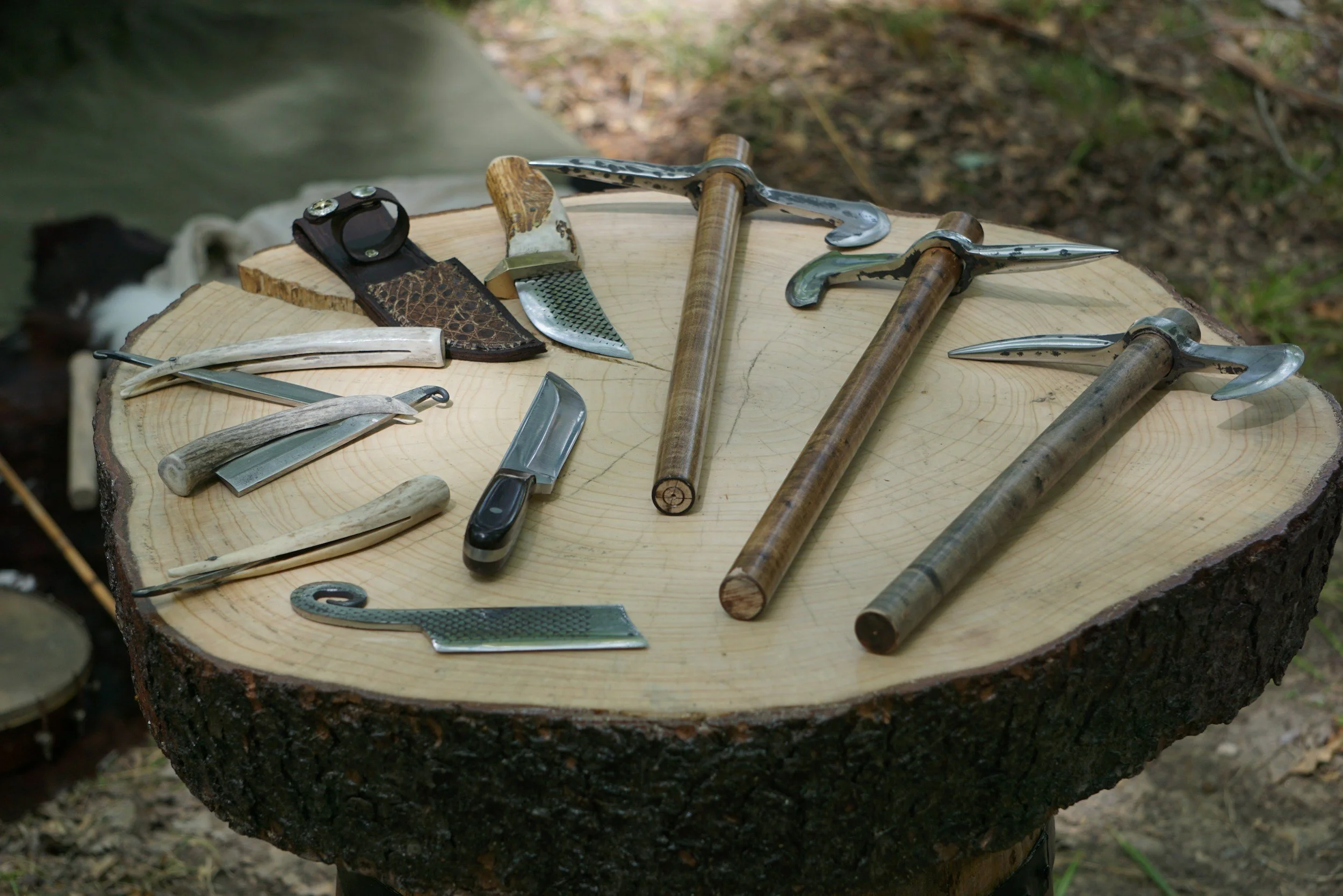 A collection of outdoor tools including three ice axes, knives, and other small cutting tools arranged on a large tree stump.