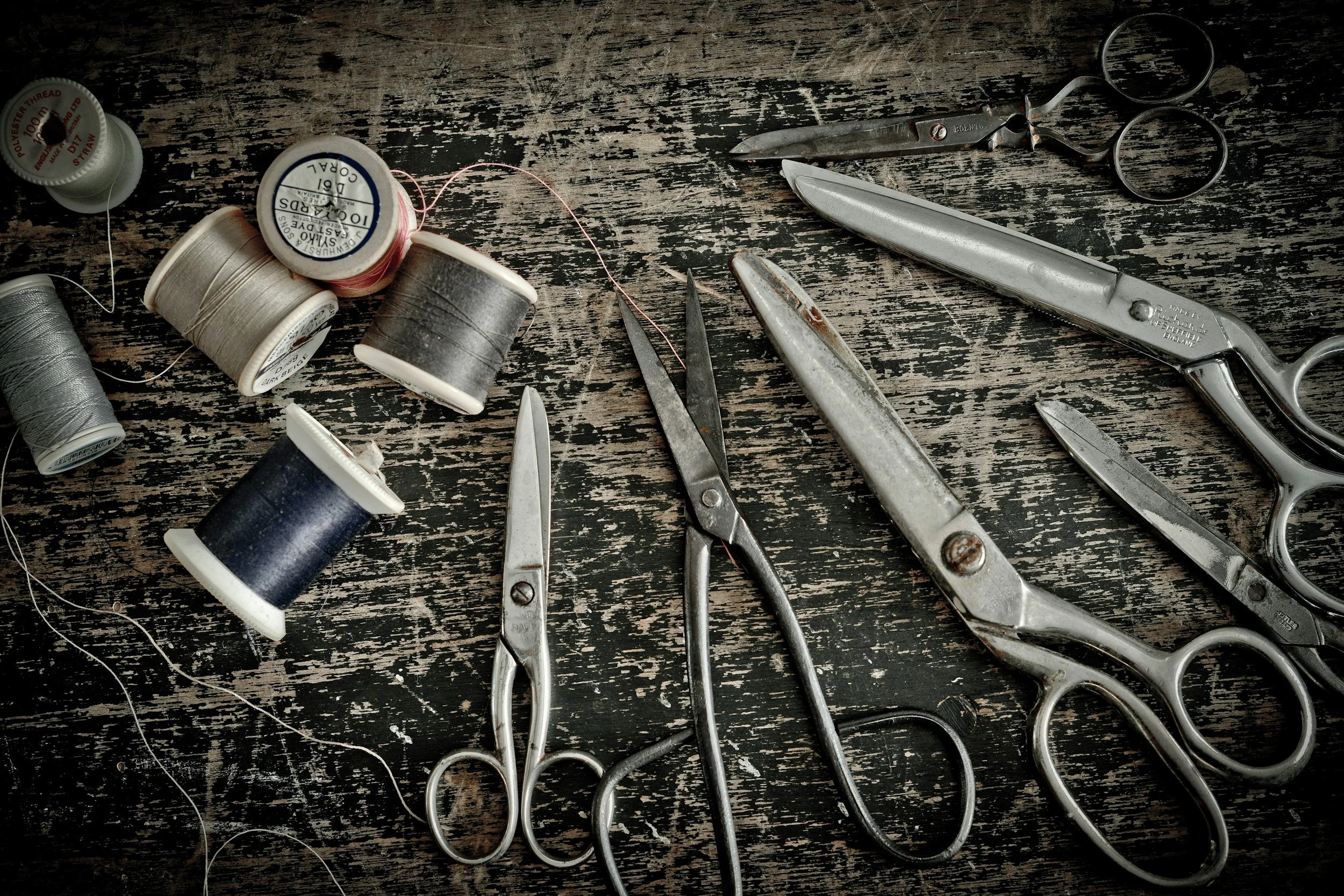 Vintage sewing tools including multiple spools of thread and several pairs of scissors on a worn wooden surface.