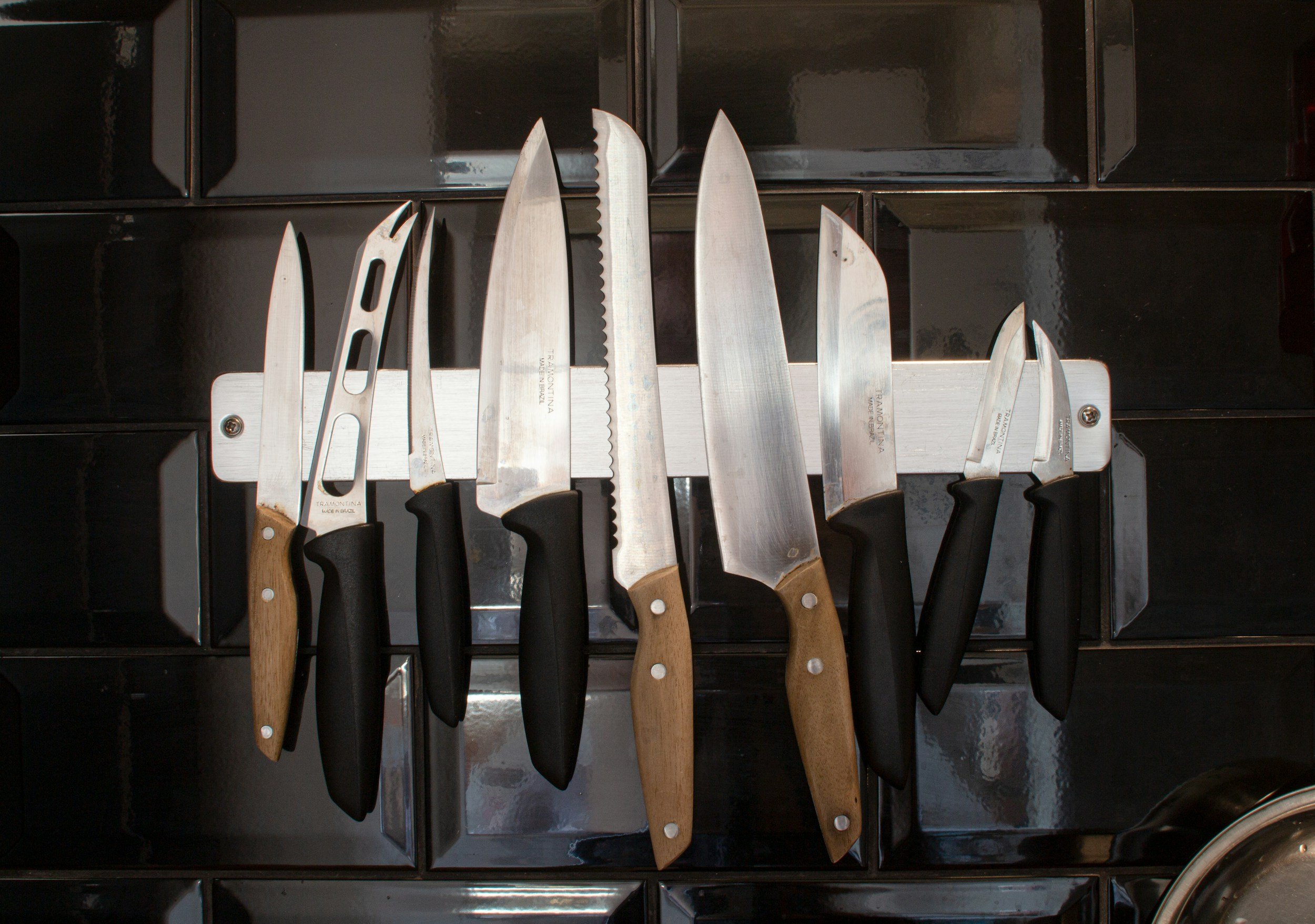 Collection of kitchen knives with black and wooden handles on a magnetic strip.