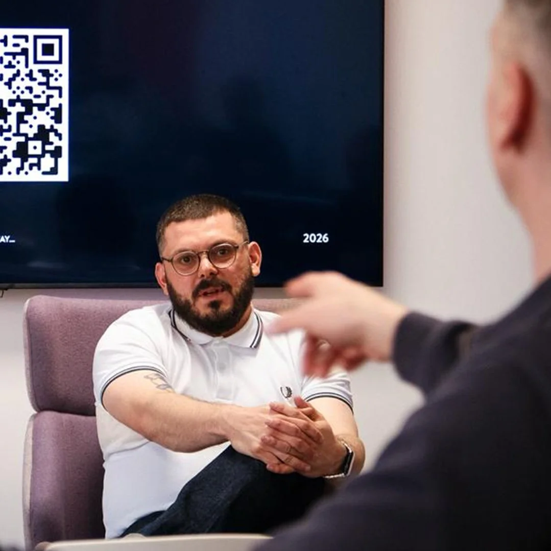 Two men in a discussion, one facing the camera wearing glasses and a white polo shirt, seated in a purple chair, the other with his back to the camera, gesturing with his hand. A screen with a QR code and the year 2026 is visible in the background.