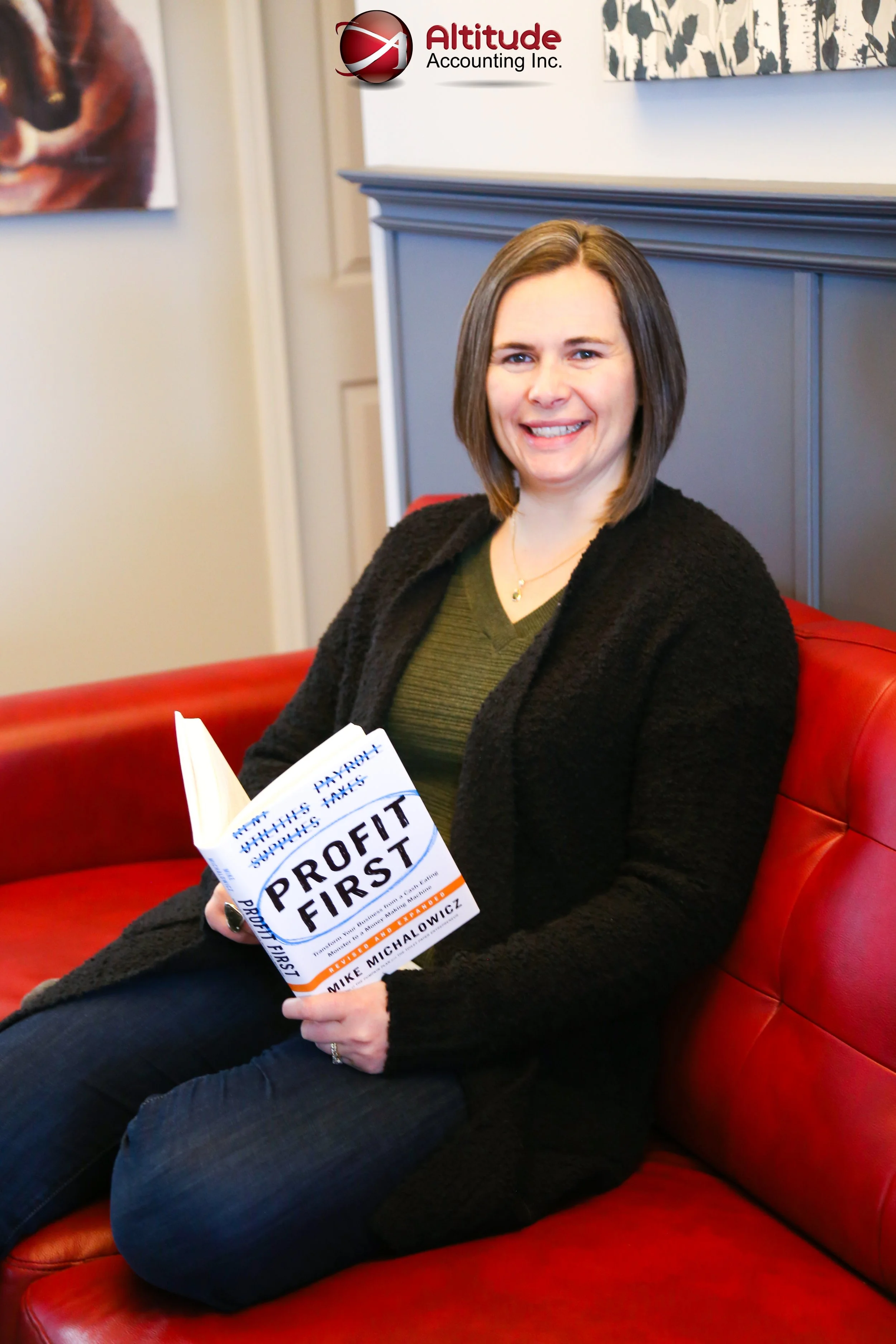 A woman sitting on a red couch, smiling and holding a book titled 'Profit First' by Mike Michalowicz, with a dark gray room divider or panel behind her.