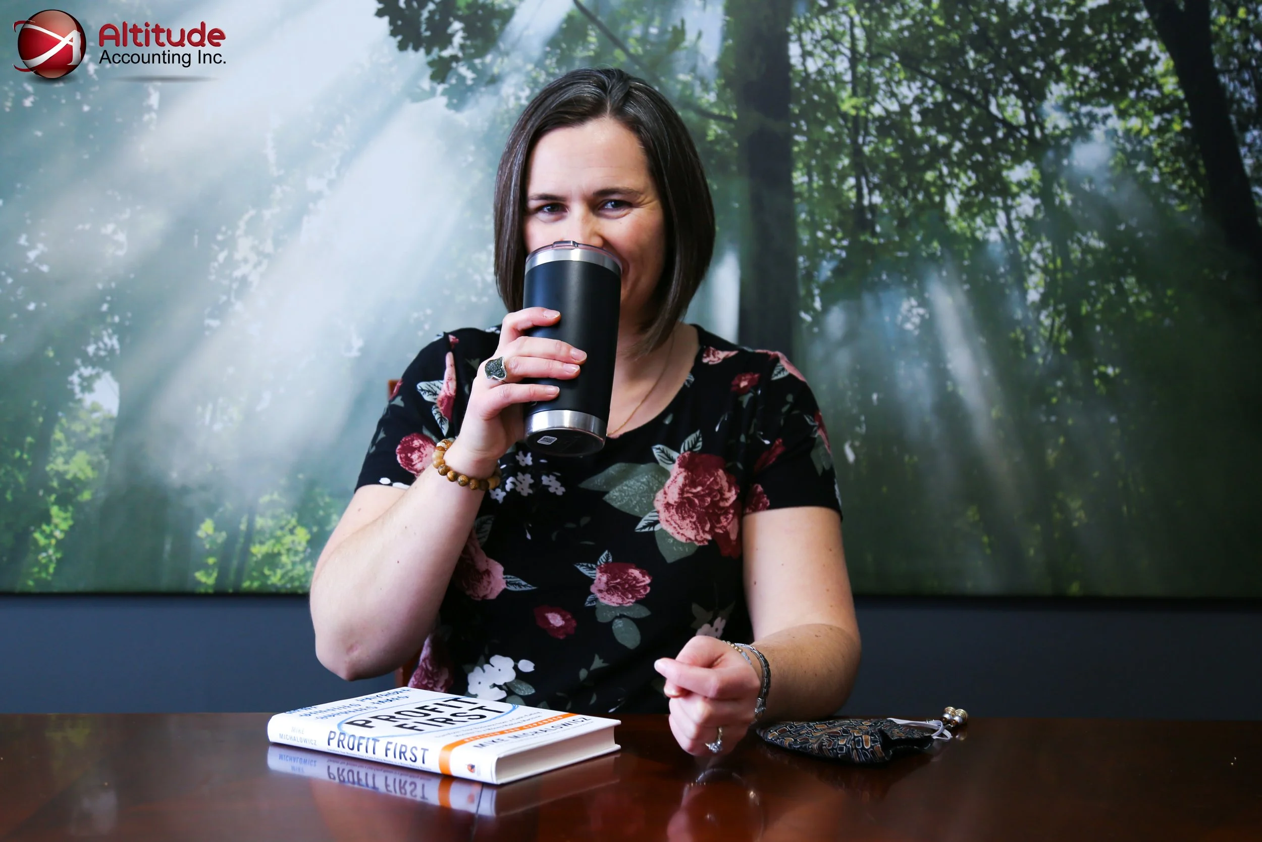 Woman with a short bob haircut drinking from a travel mug, seated at a table with a book titled "Profit First" and a small patterned bag, in front of a backdrop of a forest scene with sunlight beams.