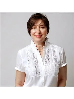 A woman with short brown hair wearing a white blouse with lace details, smiling against a plain gray background.