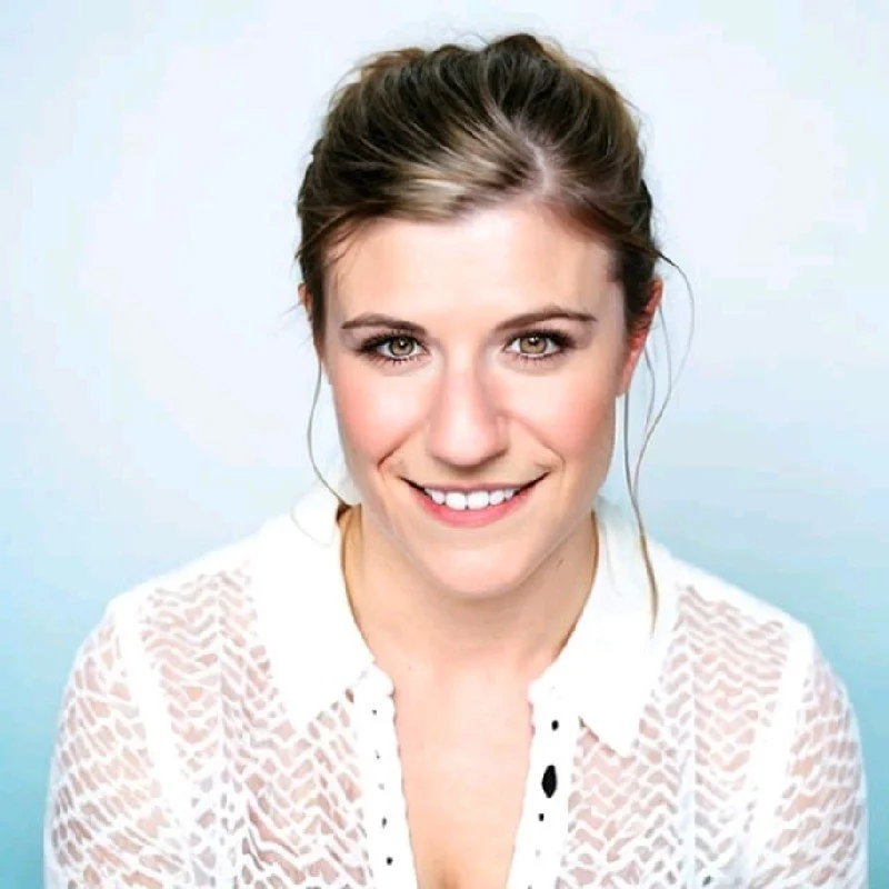 Close-up portrait of a young woman with light brown hair, smiling, wearing a white lace blouse, against a plain light blue background.