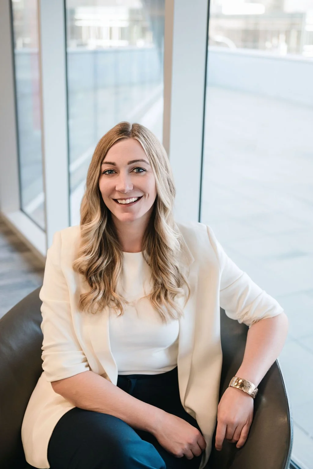 Smiling woman with long blonde hair wearing a white blazer and black pants sitting in a modern office lounge.