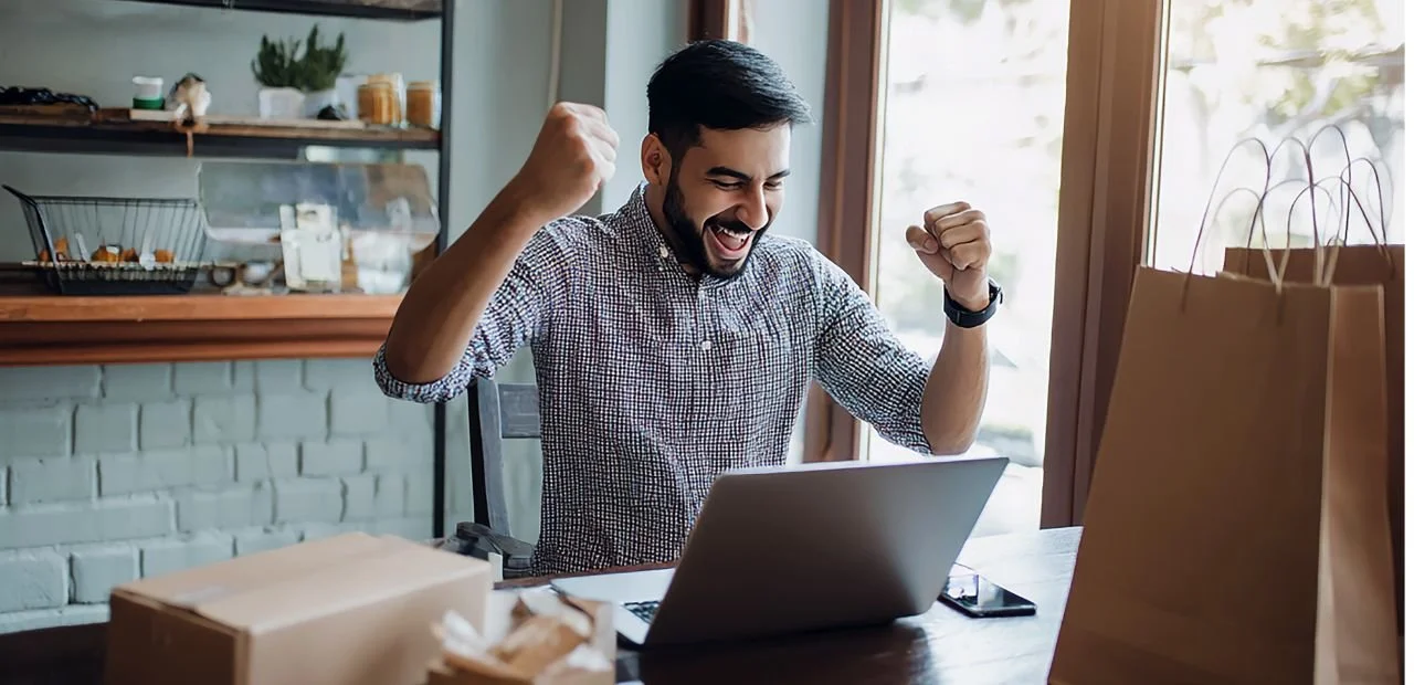 Happy man celebrating success while sitting at a table with a laptop, surrounded by shopping bags and a smartphone.