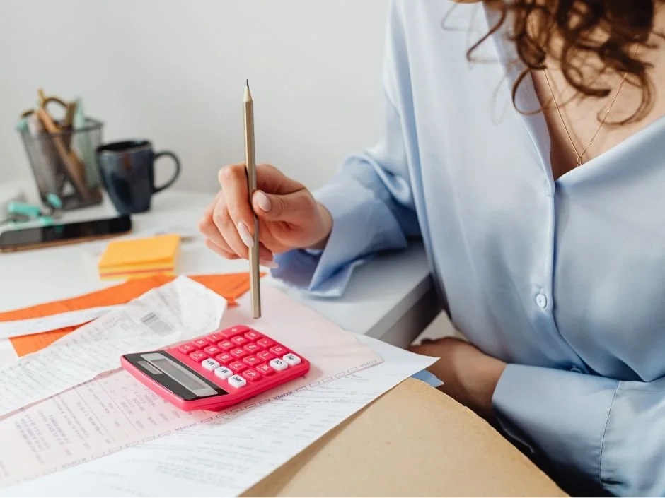 Person working at a desk with a pink calculator, documents, and a pen, in a well-lit office space.