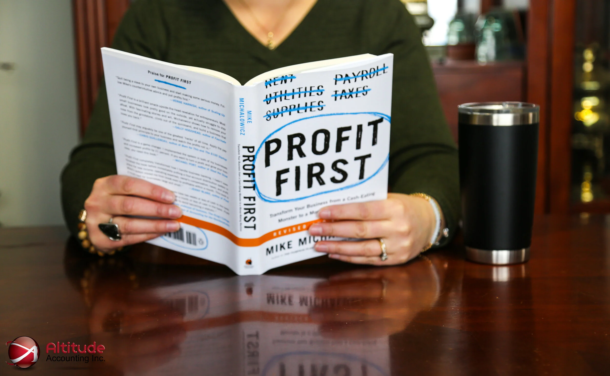 Person reading a book titled 'Profit First' by Mike Michalowicz, with a black tumbler on a wooden table.