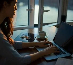 A woman working on a laptop at a table near large windows with a view of water outside.