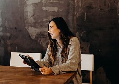 A woman sitting at a wooden table, laughing and holding a tablet, with a dark textured wall in the background.