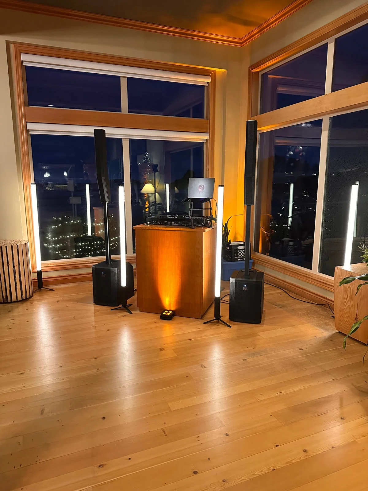Indoor space with large windows showing a nighttime cityscape, wooden floors, and a DJ setup with speakers, lights, and equipment.