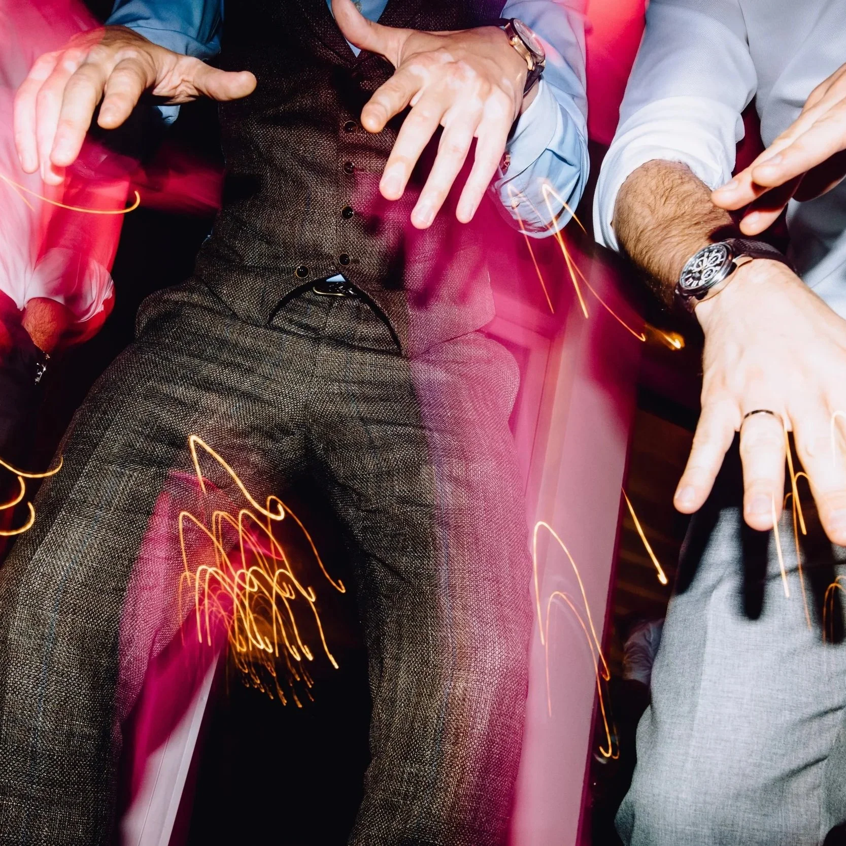 Two men in dress shirts and vests making hand gestures, with light streaks and streaks of pink light around them, suggesting a lively atmosphere.